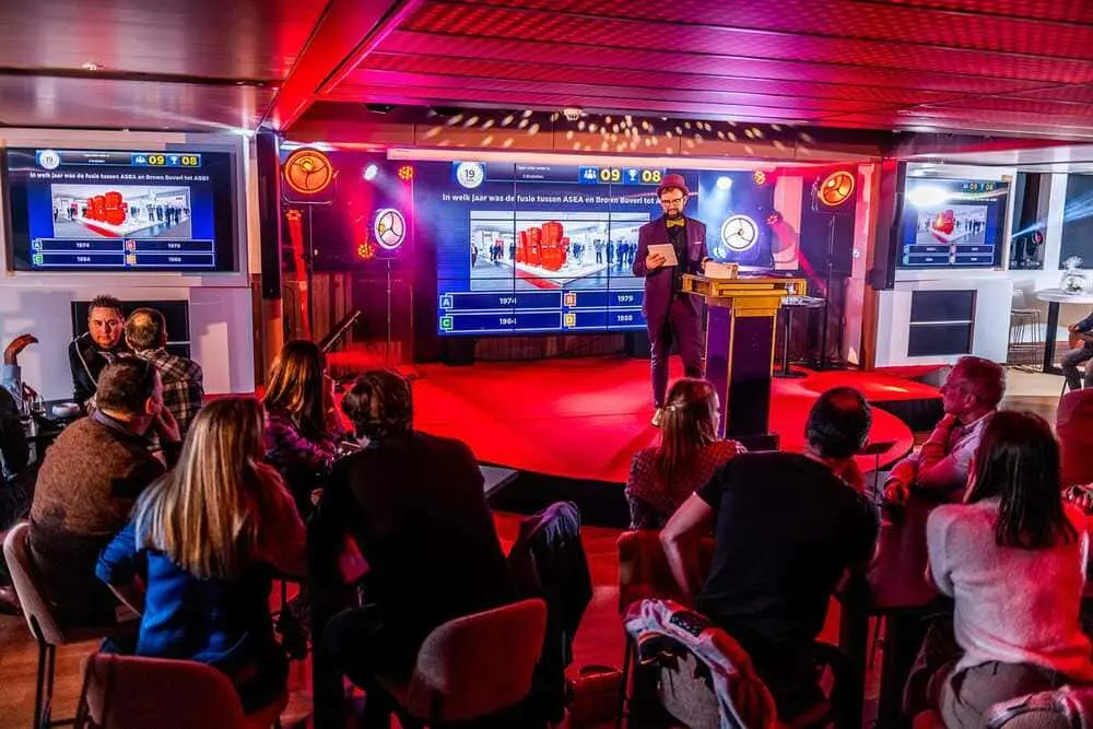 Grote zaal met meer dan 200 deelnemers aan een pubquiz met meerdere schermen