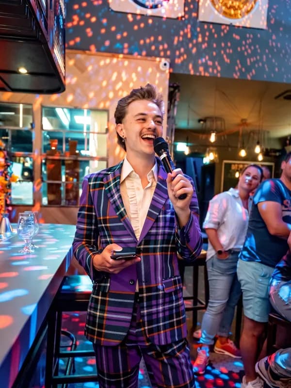 Quizmaster in paars ruit-pak Jonge quizmaster in een paars-geruit pak spreekt door een microfoon in een sfeervolle horeca- en bar-setting.