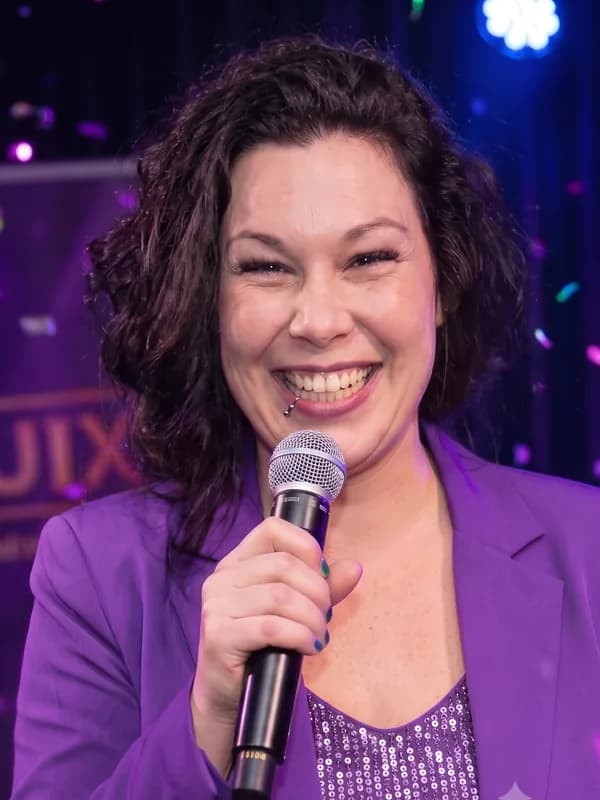Presentatrice Roxy in paars colbert Presentatrice Roxy met donkerbruin krullend haar in een paars colbert en pailletten top spreekt door een microfoon op een QuizX-avond.