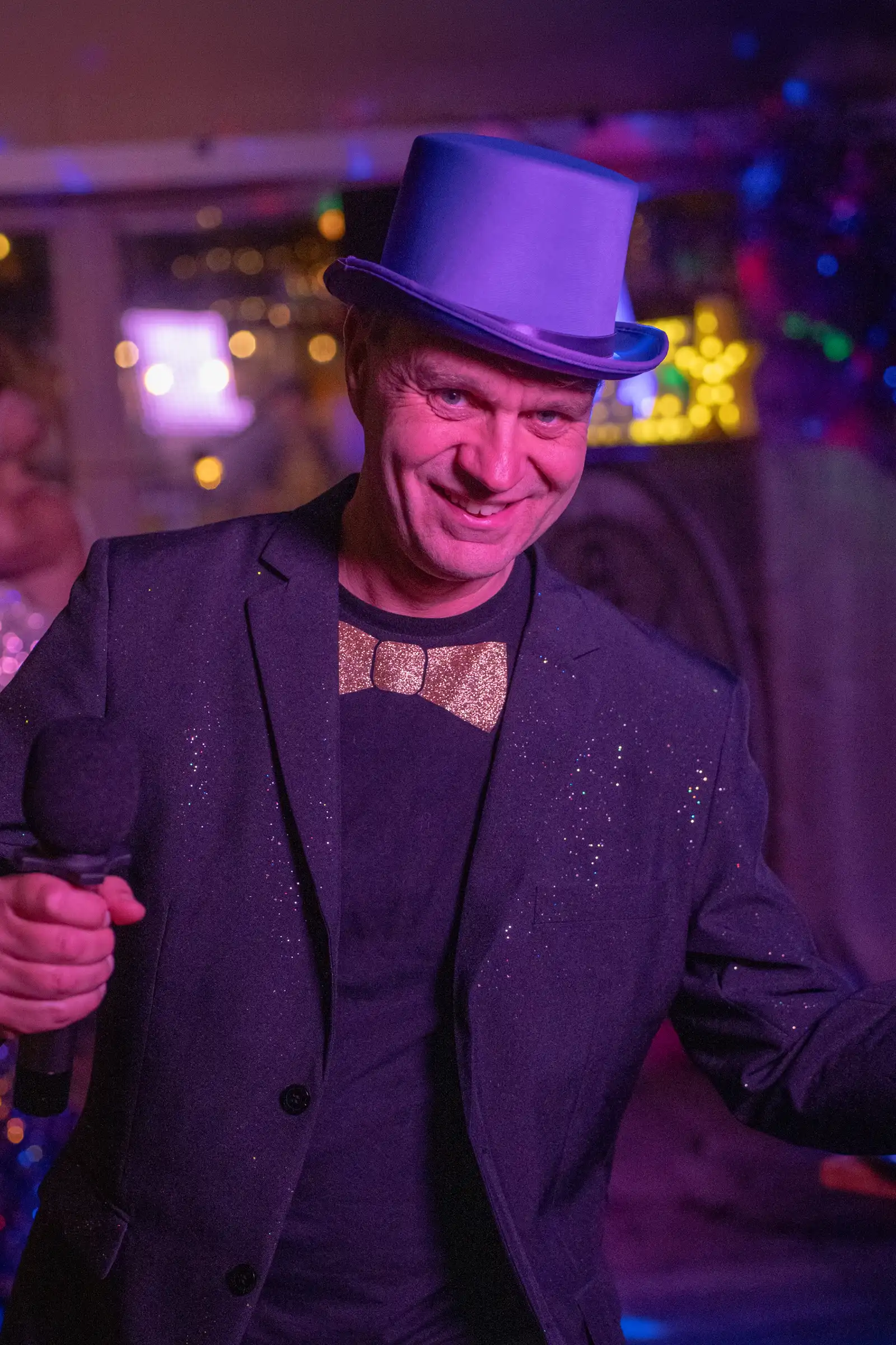 Quizmaster met paarse hoed en strik Quizmaster in een jasje met glitters, een gouden strik en paarse hoed glimlacht met een microfoon in de hand.