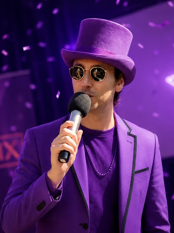 Quizmaster Kees in paars pak Quizmaster Kees met paarse hoge hoed en zonnebril in een paars pak tussen de confetti tijdens een QuizX-avond.