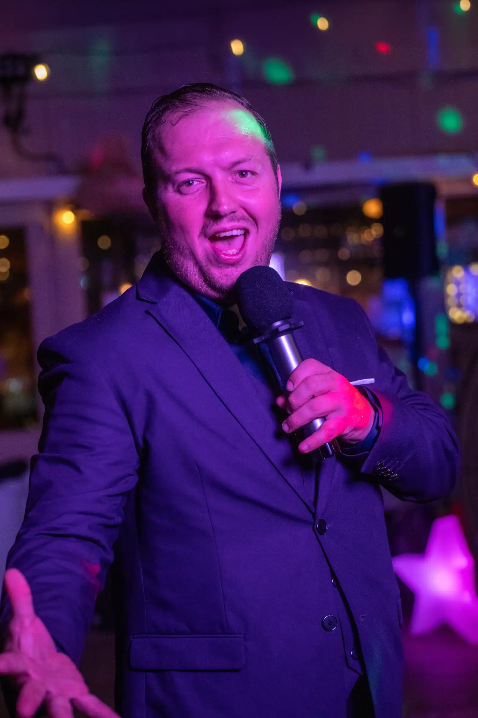 Quizmaster Lenio met blauwe hoed Lachende quizmaster Lenio met een blauwe hoed spreekt enthousiast door een microfoon in een sfeervol verlichte zaal.