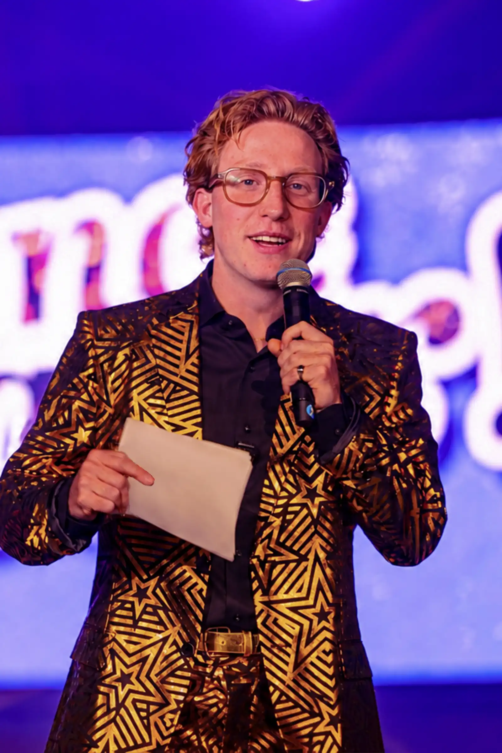 Quizmaster Bram Goud Pak Microfoon Quizmaster Bram in een opvallend goud pak met sterrenpatroon spreekt door de microfoon met kaartjes in zijn hand.