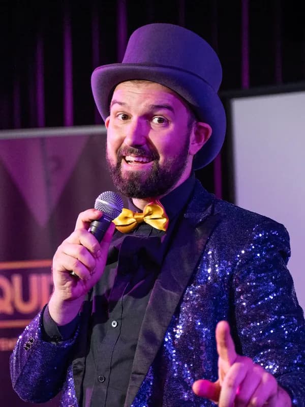 Quizmaster Liam in glittercolbert Quizmaster Liam met een hoed, strik en blauw glittercolbert spreekt door de microfoon tijdens een QuizX evenement.