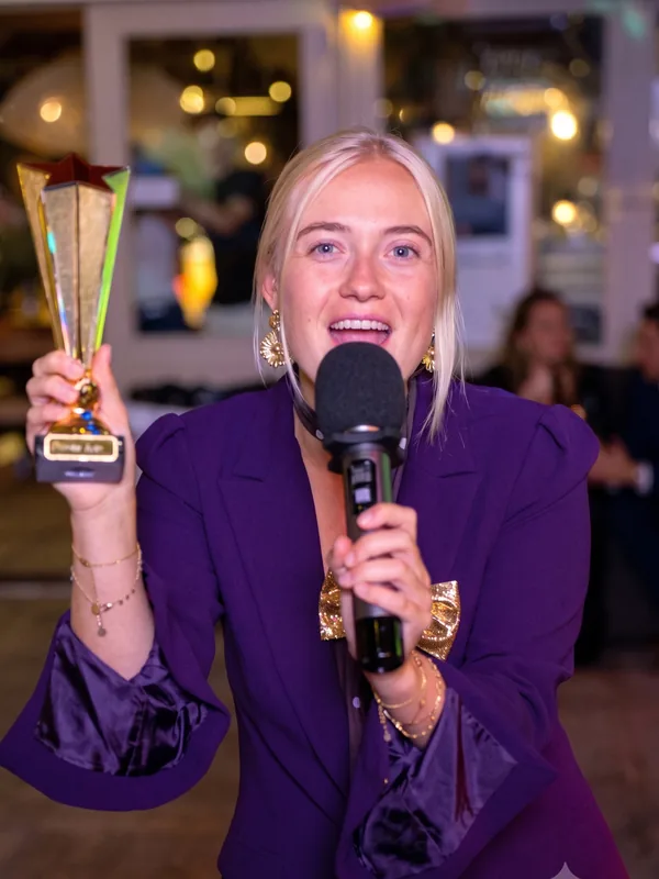 Presentatrice met gouden trofee Blonde presentatrice in een paars colbert houdt een microfoon en een gouden trofee vast tijdens een QuizX-quizshow.