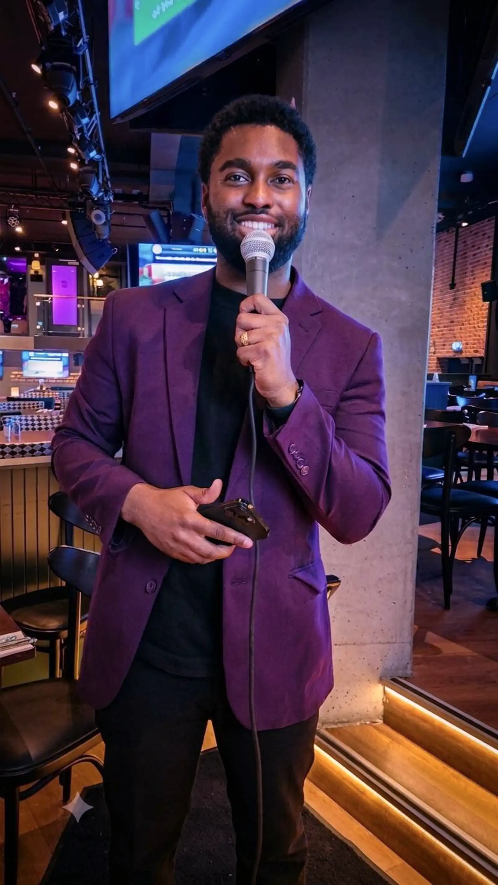 Quizmaster Paars Colbert Microfoon Horeca Lachende quizmaster in een paars colbert houdt een microfoon en smartphone vast in een sfeervolle horecagelegenheid.