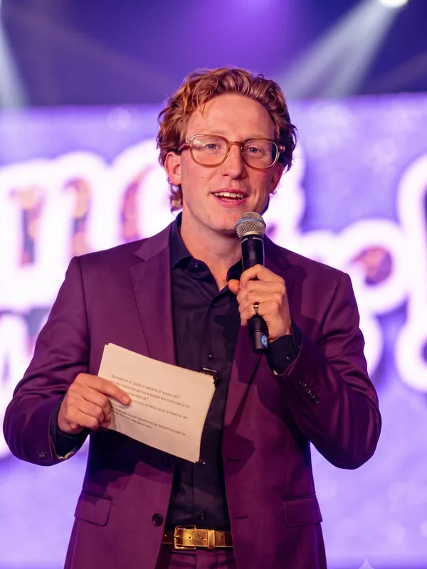 Quizmaster Bram in goud pak Quizmaster Bram in een opvallend goud pak met sterrenpatroon spreekt door de microfoon met kaartjes in zijn hand.