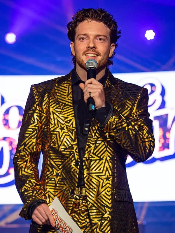 Presentator Desley in goud sterrenpak Jonge presentator Desley in een goud sterrenpak spreekt door een microfoon op het podium tijdens een QuizX-show.
