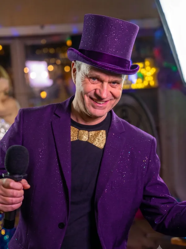 Quizmaster Peter met glitterhoed Grijzende quizmaster Peter met een paarse glitter-hoge-hoed, gouden strik en paars colbert tijdens een QuizX-show.