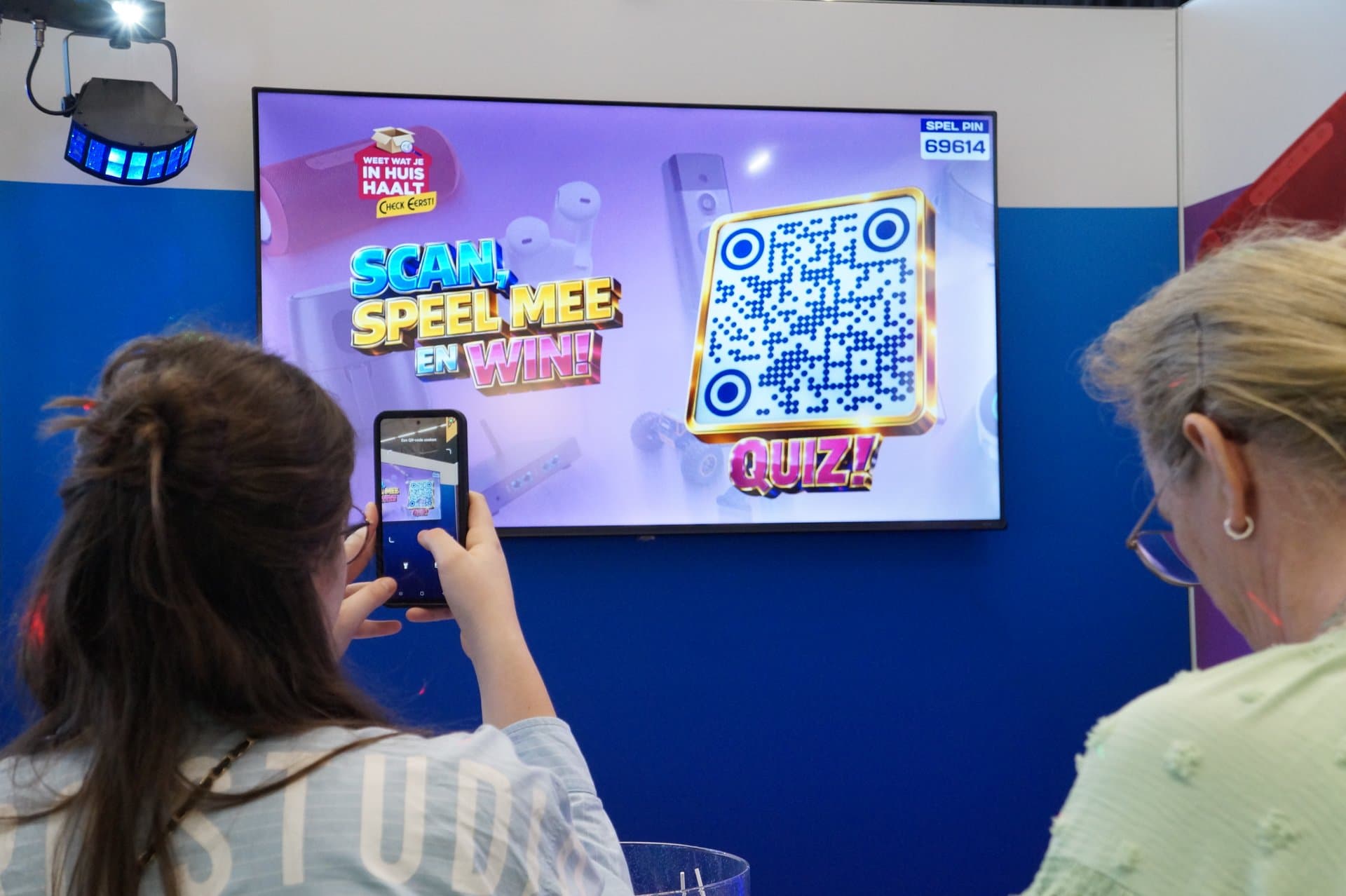 Twee deelnemers kijken naar een groot scherm met een QR-code voor een quiz. Eén persoon scant de QR-code met een smartphone, terwijl de ander toekijkt.
