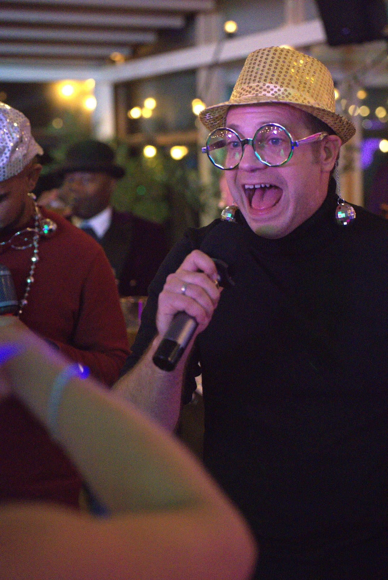 Muziekbingo meezingen met glitters Gast met gouden paillettenhoed en discobal-oorbellen zingt uit volle borst mee tijdens een muziekbingo, midden tussen de andere feestgangers.