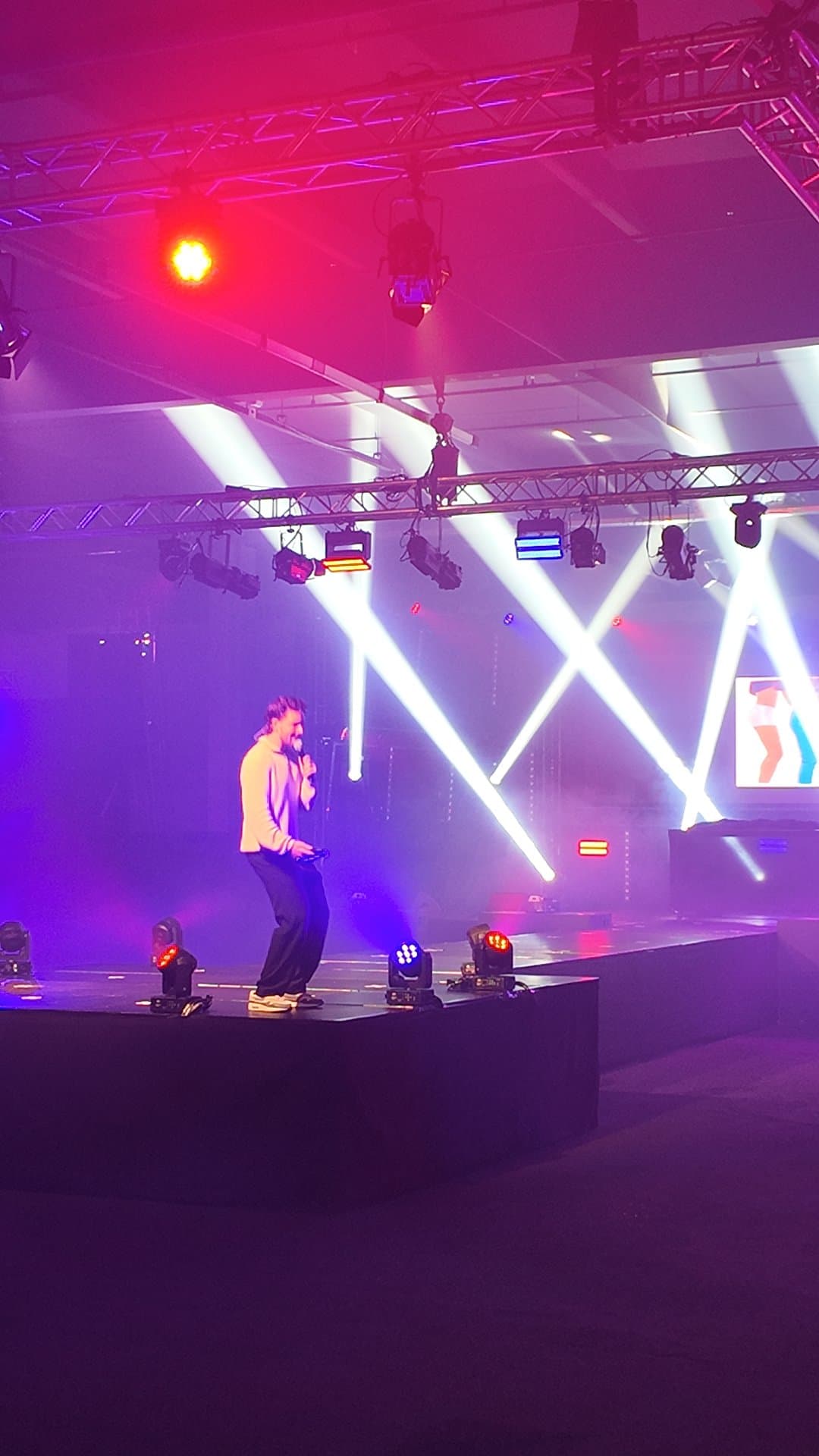 Muziekbingo energie op het podium Energieke quizmaster staat midden op het podium van een muziekbingo, omringd door dynamisch paars en rood showlicht dat de zaal opzweept.