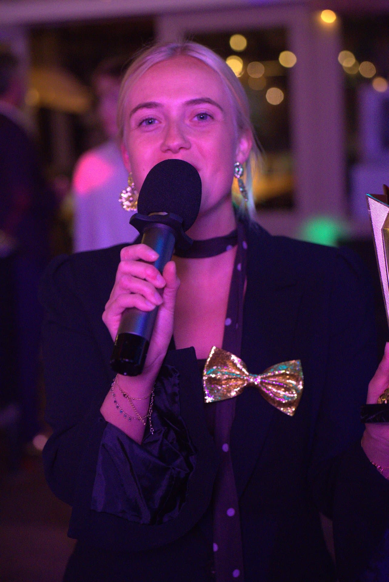Muziekbingo presentatrice paars licht Presentatrice met gouden strik kijkt recht in de camera tijdens een muziekbingo, met de microfoon in de hand en paars podiumlicht om haar heen.