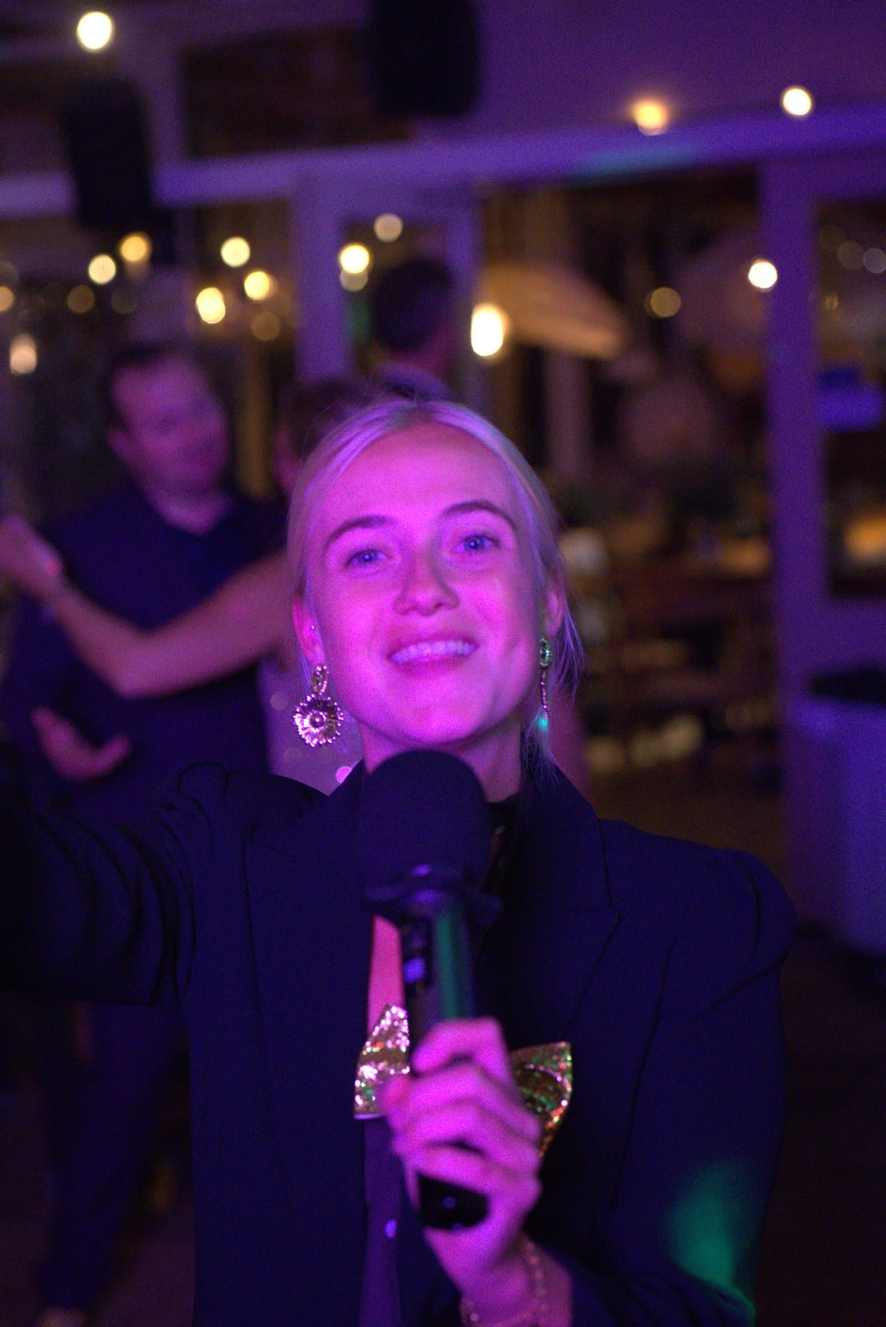Muziekbingo live zangeres Zangeres in zwarte blazer treedt op tijdens een muziekbingo en zingt lachend in de microfoon met sfeervolle lichtjes op de achtergrond.