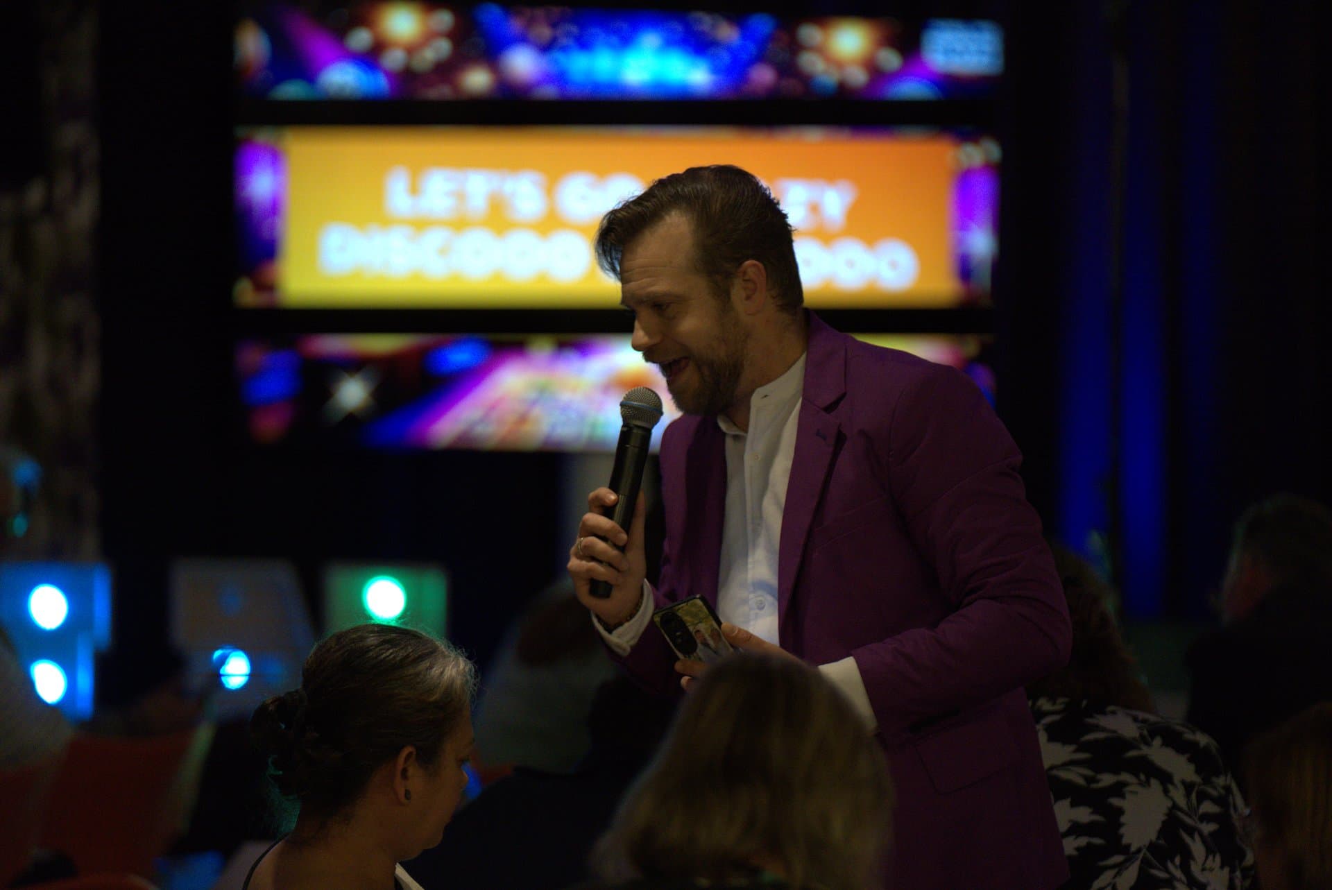 Muziekbingo quizmaster show Quizmaster in paars jasje zweept het publiek op tijdens een muziekbingo, met de tekst LETS GO DISCO op het scherm achter hem.