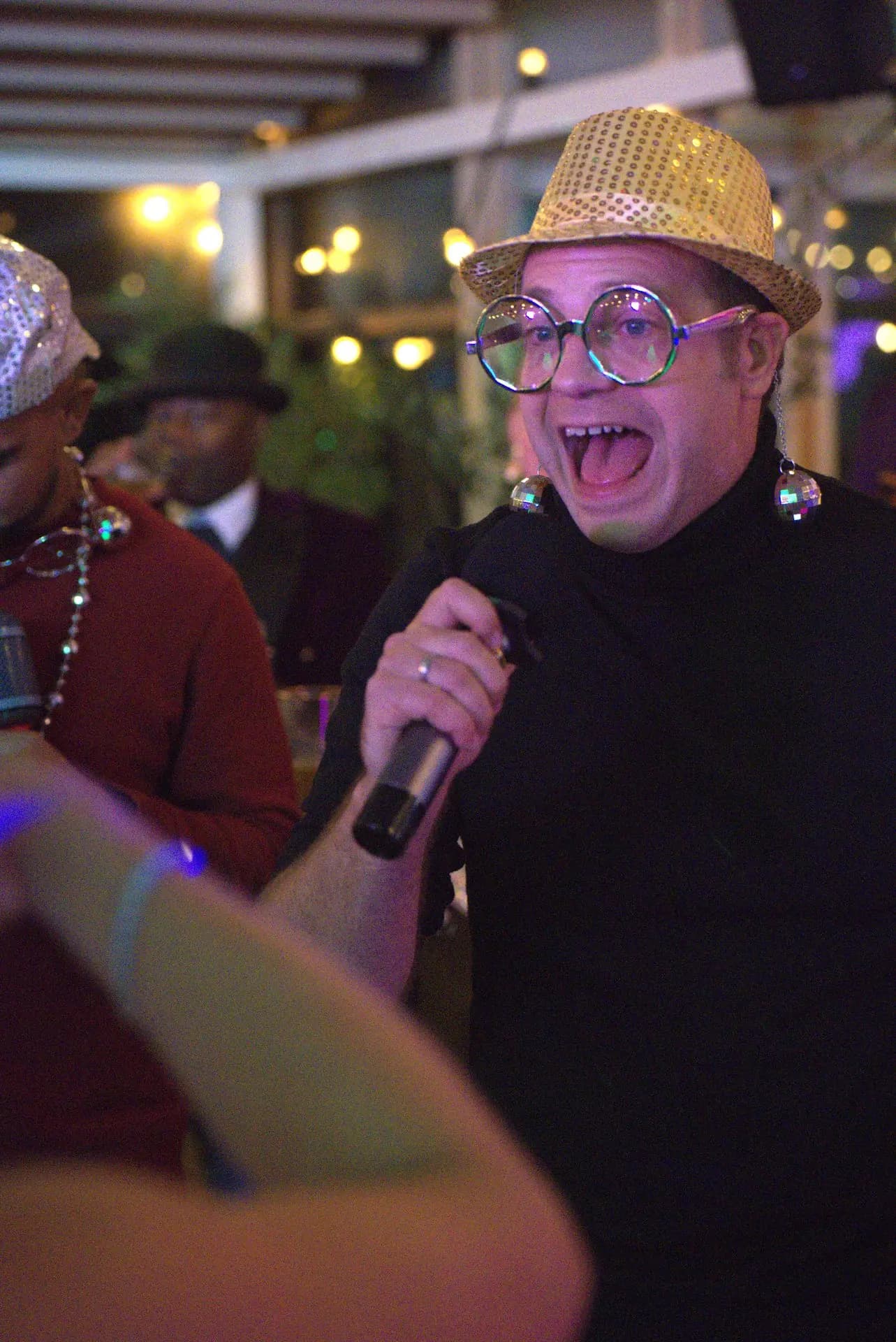 Gast met gouden paillettenhoed en discobal-oorbellen zingt uit volle borst mee tijdens een pubquiz op locatie, midden tussen de andere feestgangers.