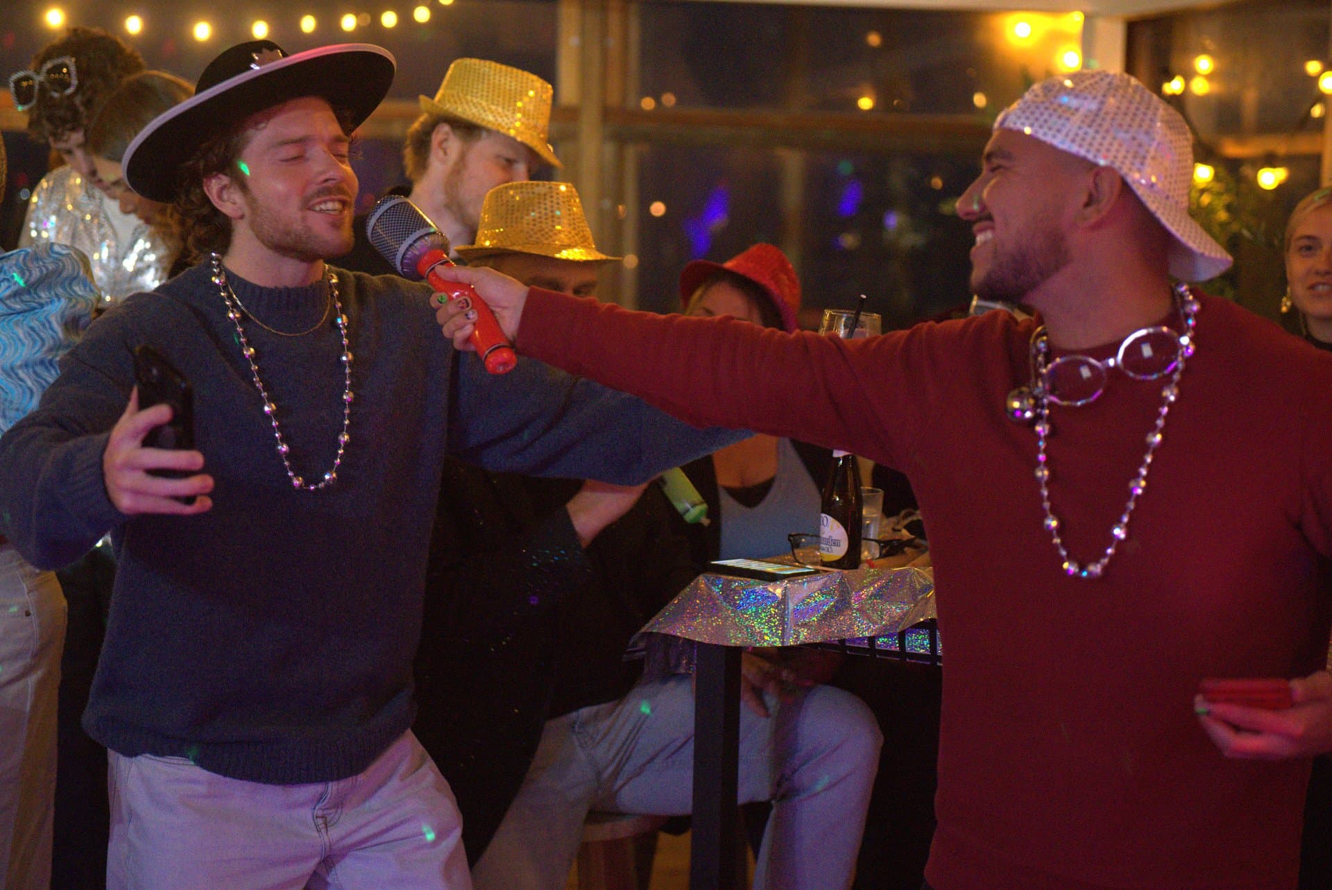 Pubquiz voor bedrijven karaoke afterparty Twee deelnemers zingen samen karaoke tijdens de afterparty van een pubquiz voor bedrijven, omringd door gasten met feesthoeden en accessoires.