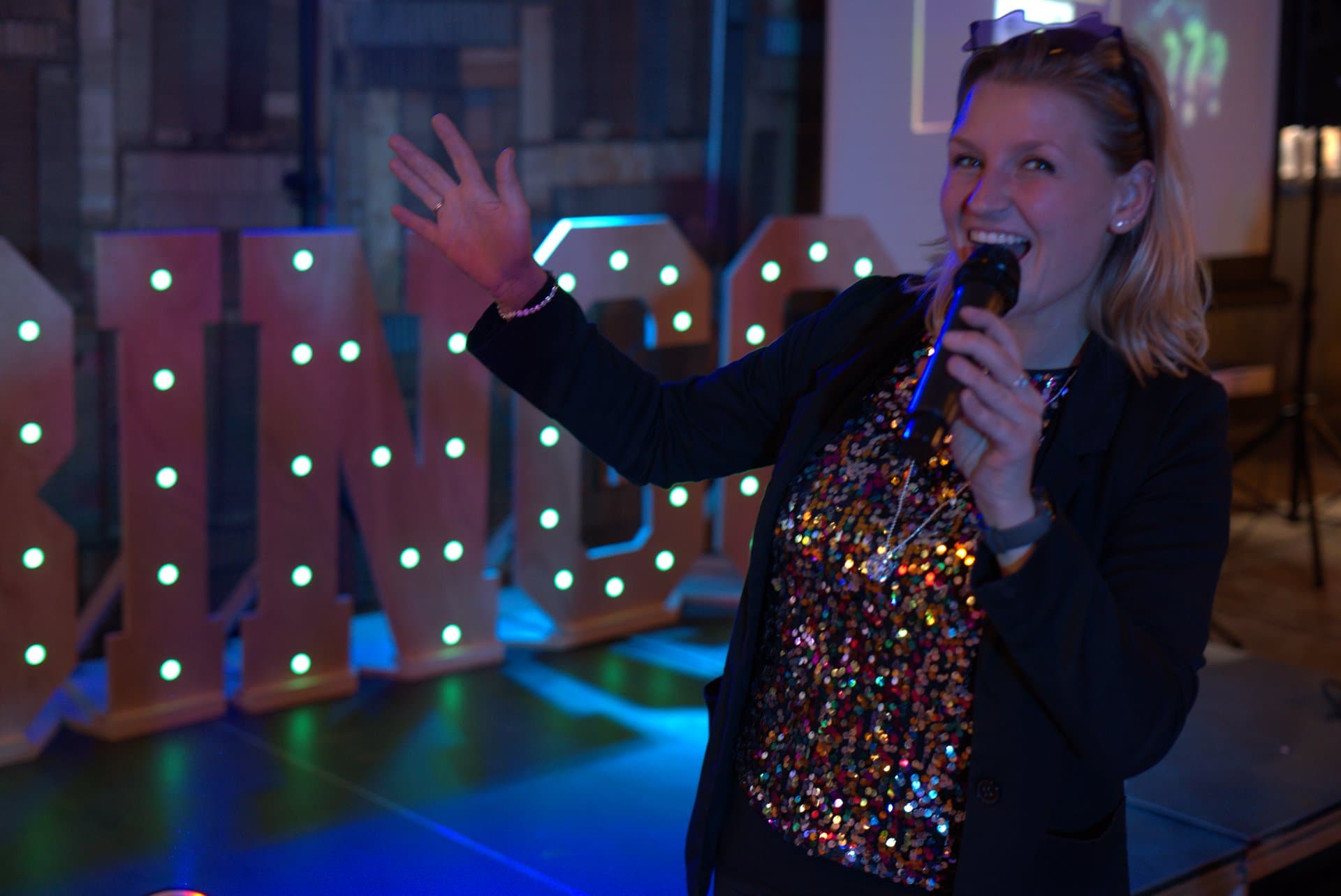 Pubquiz voor bedrijven quizmaster met microfoon Lachende quizmaster met microfoon staat voor verlichte QUIZ-letters, klaar om een pubquiz voor bedrijven met energie aan elkaar te presenteren.