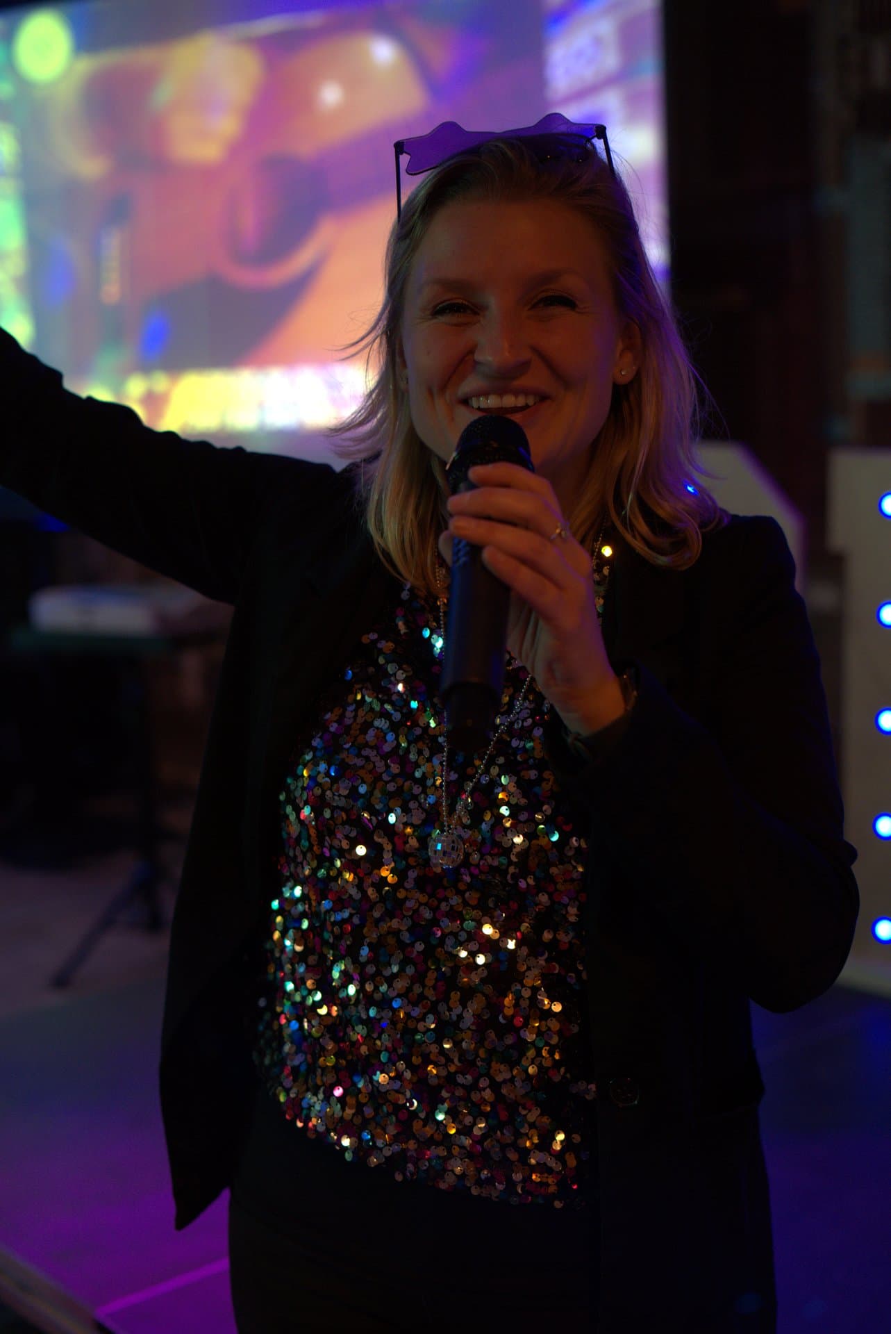 Pubquiz voor bedrijven quizmaster op podium Quizmaster met glimmende top en paarse zonnebril staat stralend op het podium van een pubquiz voor bedrijven, met kleurrijke lichten achter haar.