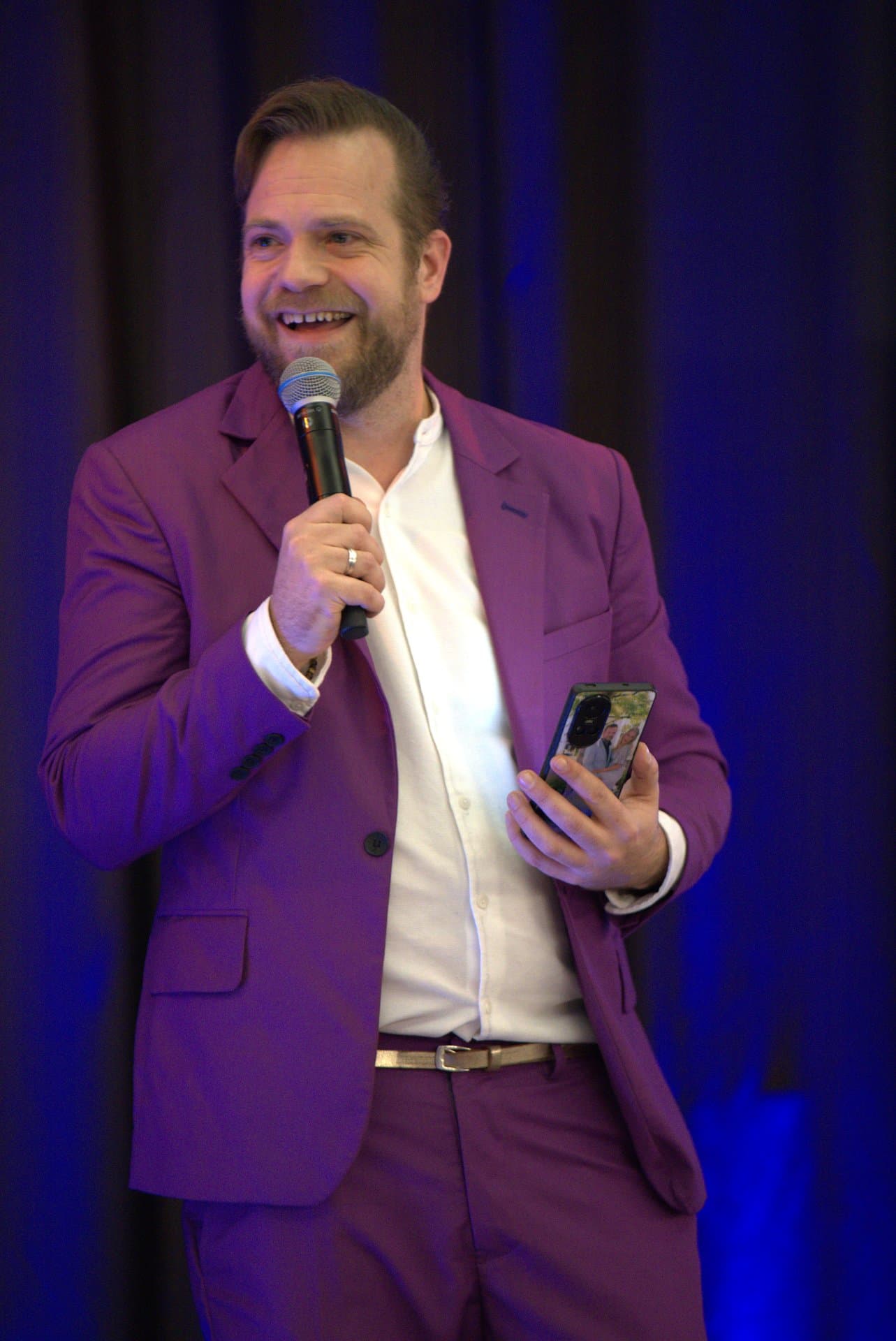 Pubquiz voor bedrijven fotoronde uitleg Quizmaster in paars pak toont een foto op zijn telefoon tijdens een pubquiz voor bedrijven en praat de zaal door de fotoronde heen, microfoon in de andere hand.