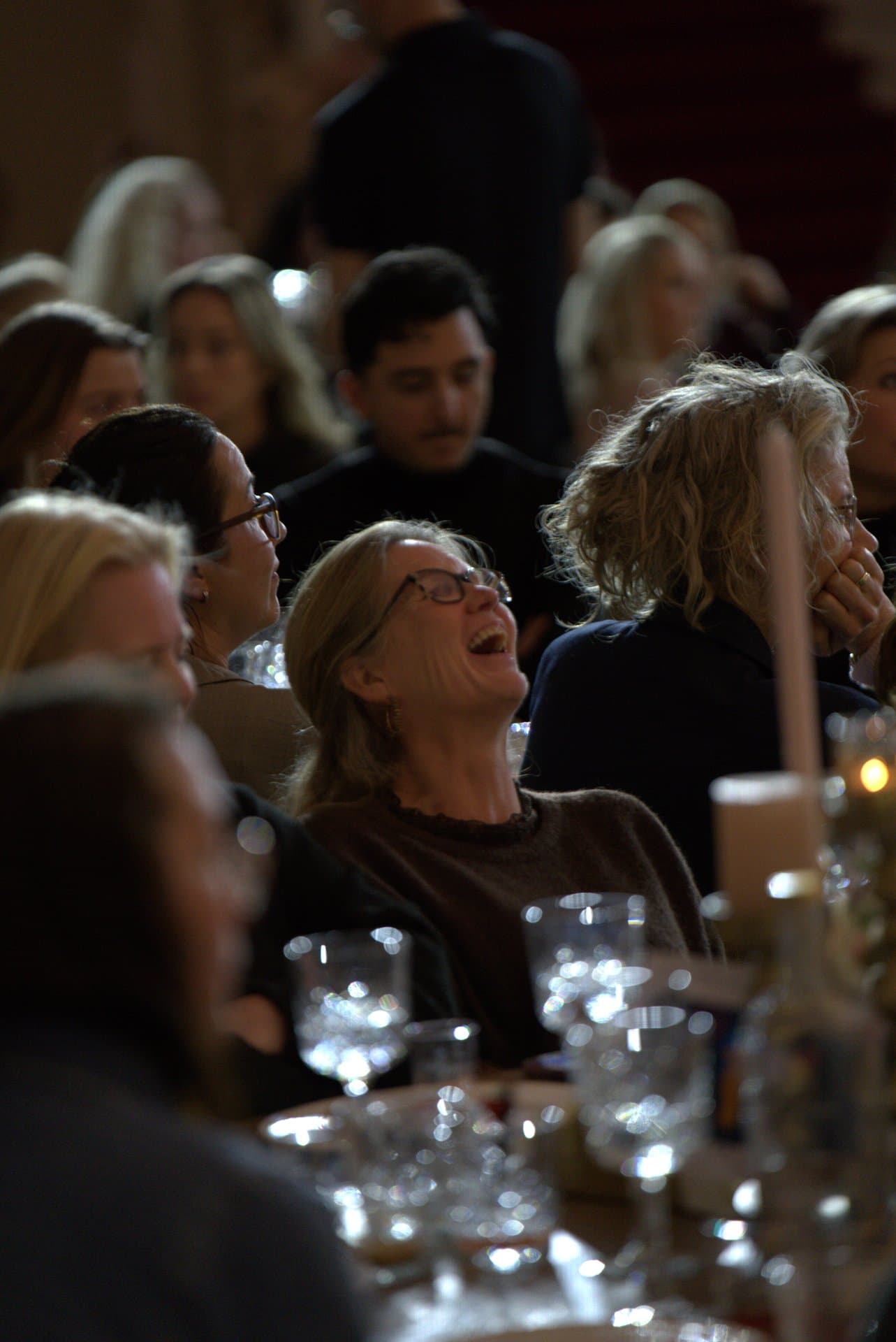 Pubquiz voor bedrijven hardop lachen Deelneemster met bril lacht hardop aan tafel tijdens een pubquiz voor bedrijven, tussen glimmende glazen en teamgenoten in opperbeste feeststemming.