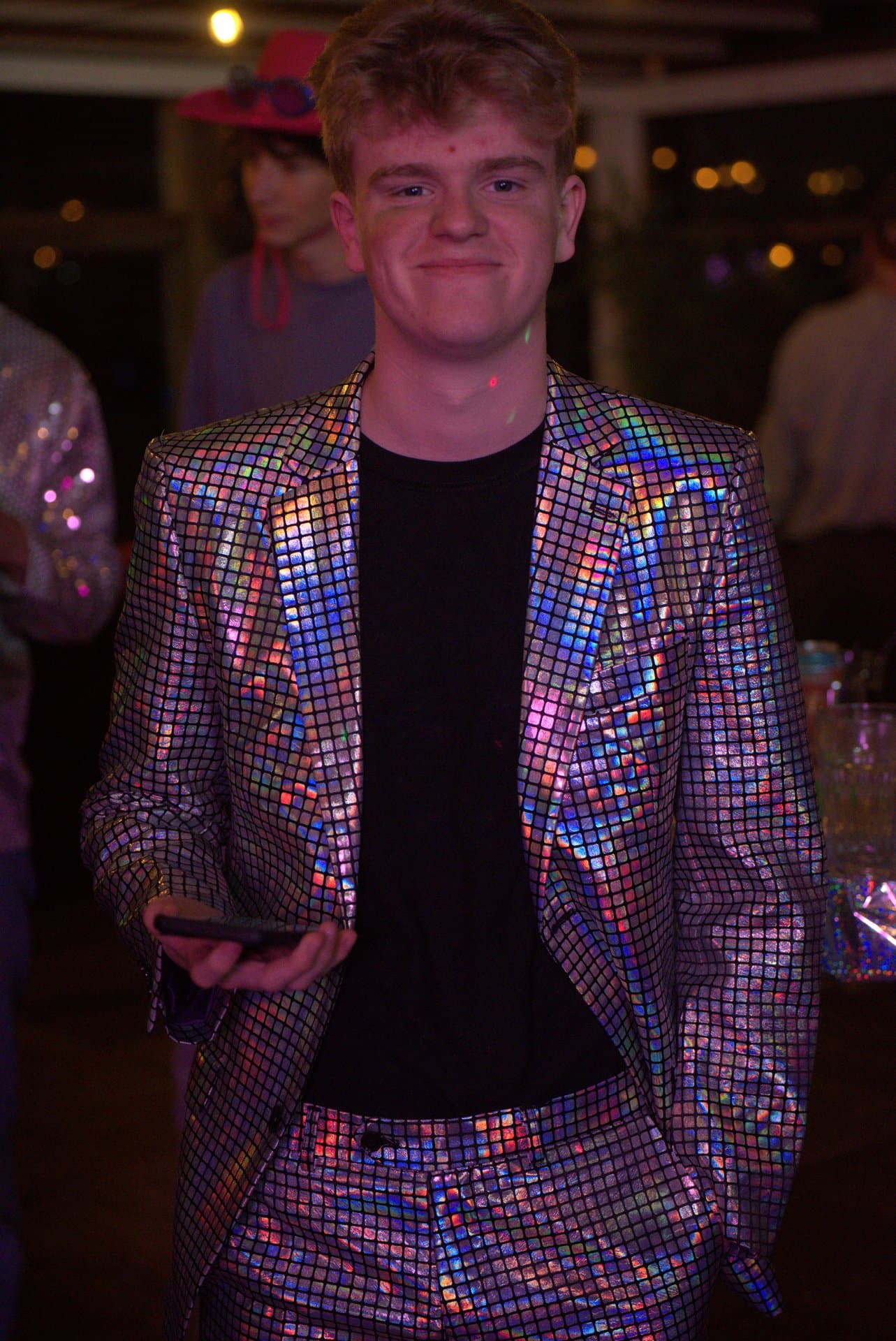Disco bingo - Een jonge man in een glimmend, spiegelend pak glimlacht naar de camera tijdens een feestelijk evenement. De...