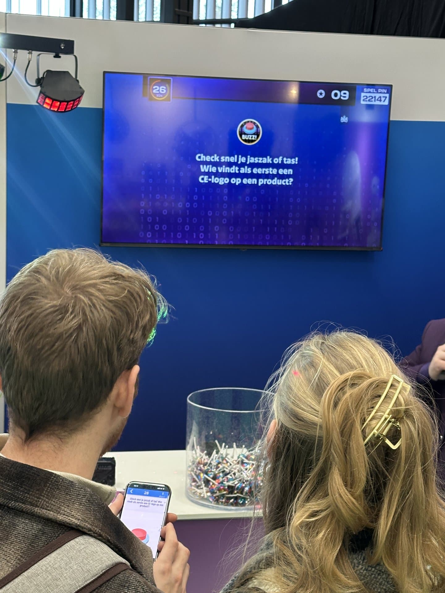 Pubquiz met quizmaster - Twee deelnemers kijken naar een groot scherm tijdens een quiz, waarbij de vraag is wie als eerste een CE-logo...