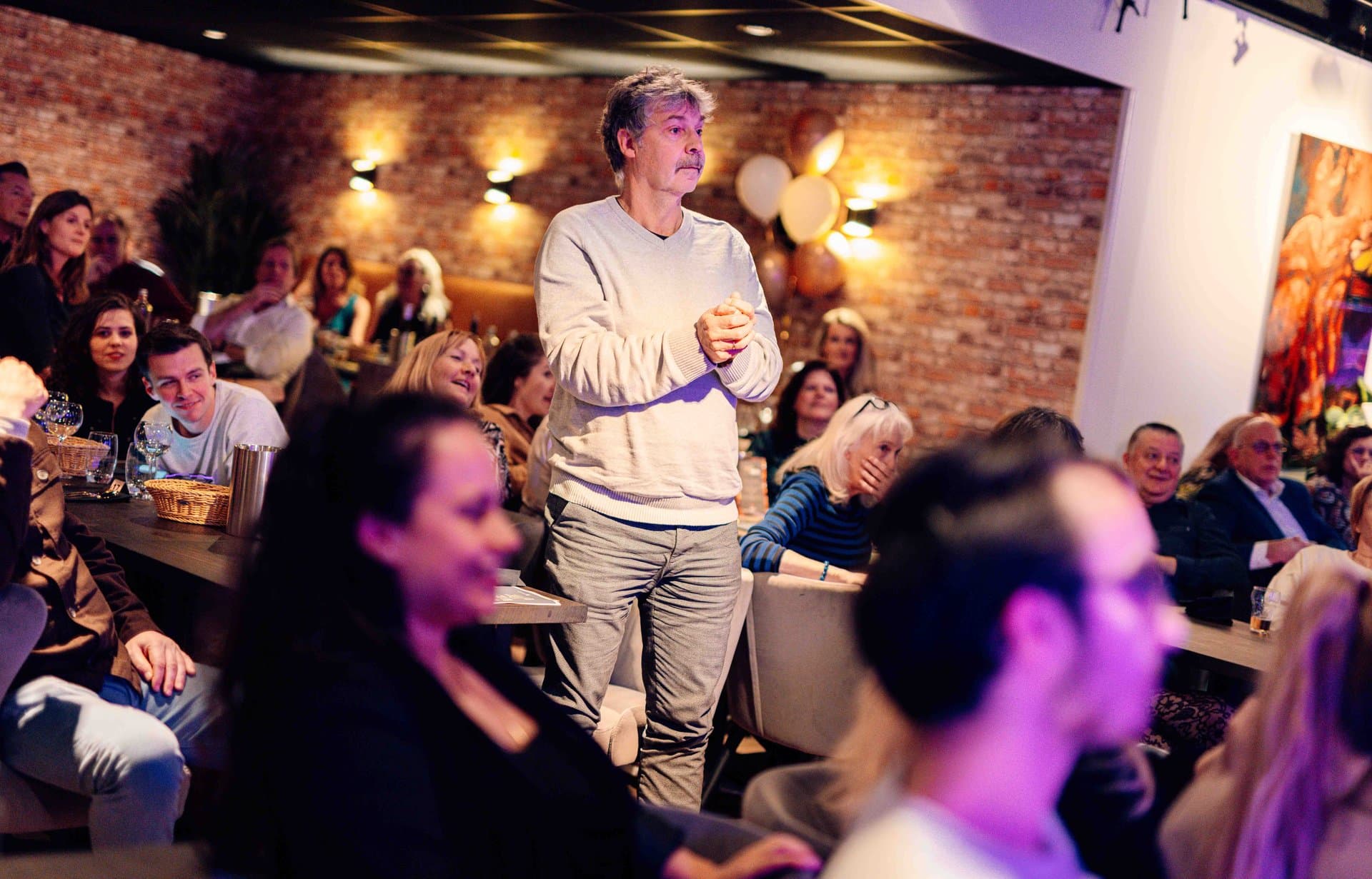 Pubquiz bedrijfsuitje - Een quizmaster staat centraal en spreekt een aandachtig publiek toe in een sfeervolle ruimte met bakstenen...