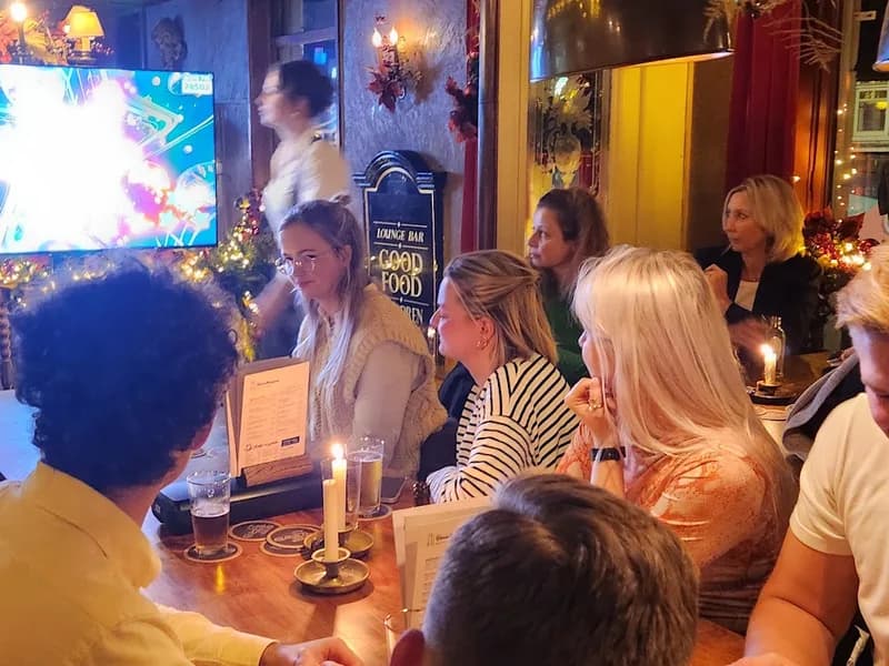 Pubquiz bedrijfsuitje - Een quizmaster staat centraal en spreekt een aandachtig publiek toe in een sfeervolle ruimte met bakstenen...