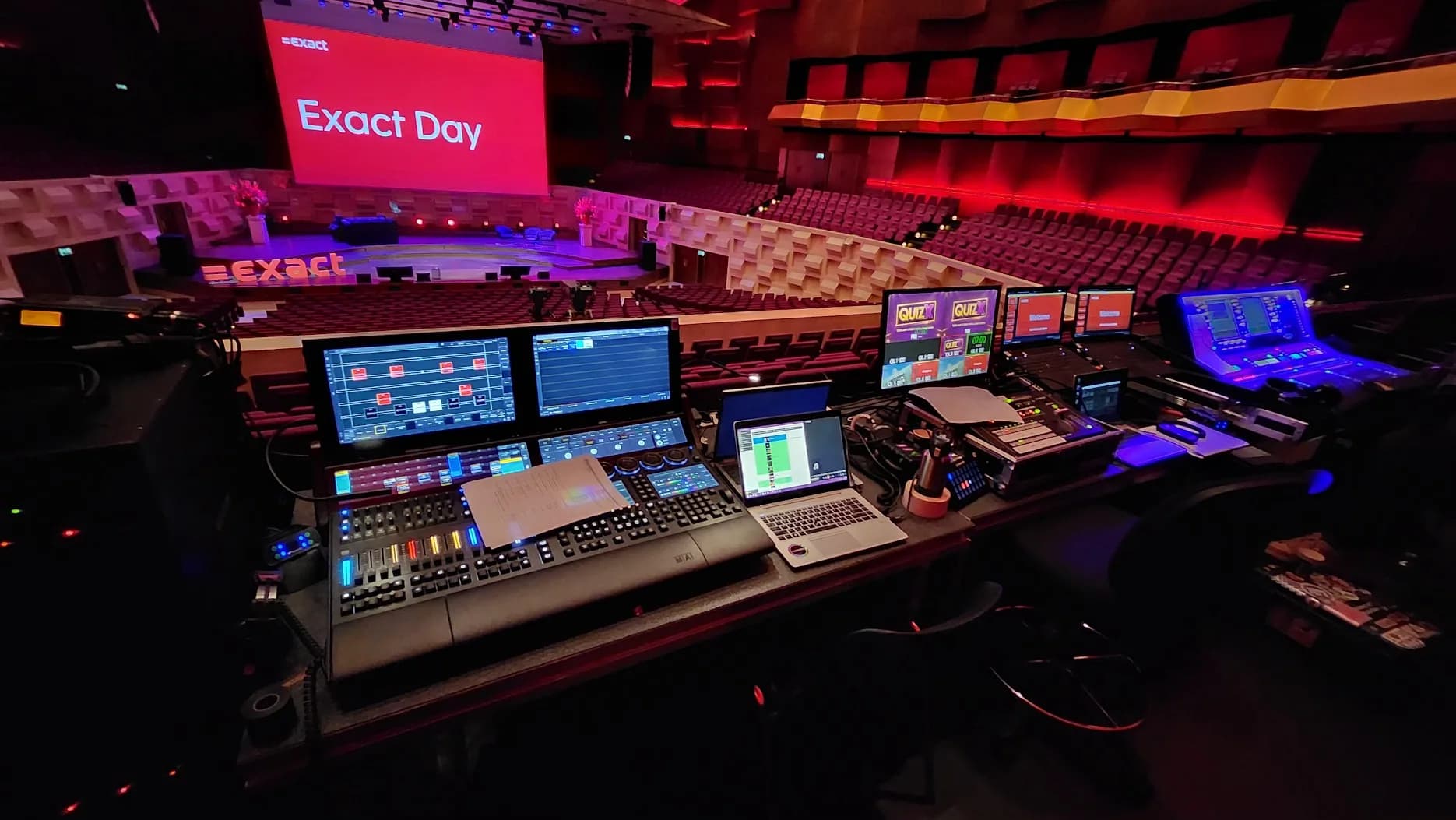 Regietafel achter in de Grote Zaal van De Doelen met lichttafel, geluidstafel en de quizlaptop van QuizX, het podium met Exact Day op het scherm op de achtergrond