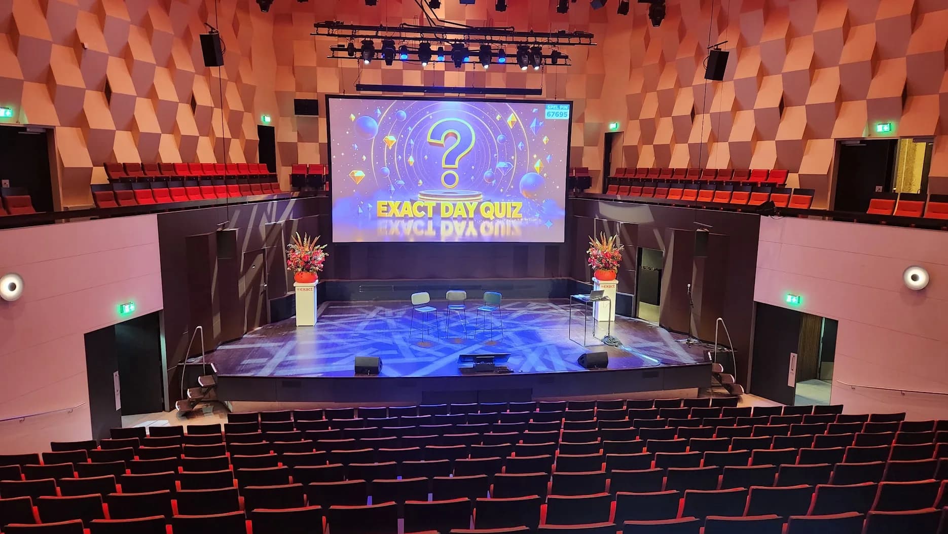 Een van de kleinere zalen van De Doelen met het titelscherm Exact Day Quiz en de spelpin op het scherm, drie krukken op het podium