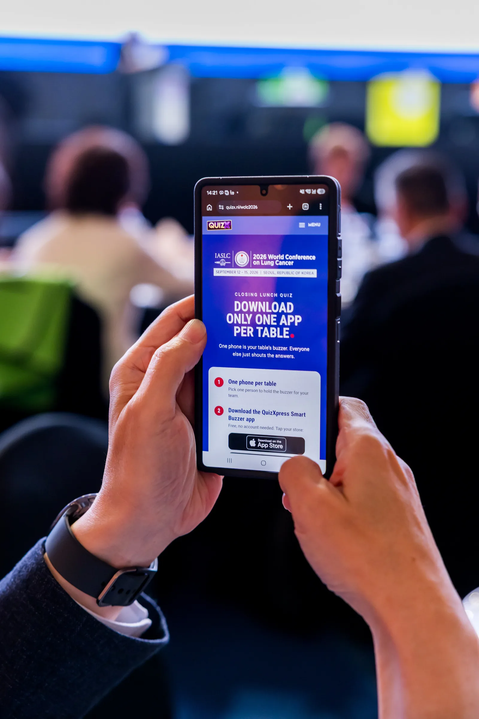 Twee handen houden een telefoon vast met de WCLC 2026-startpagina van de buzzer-app, de zaal op de achtergrond