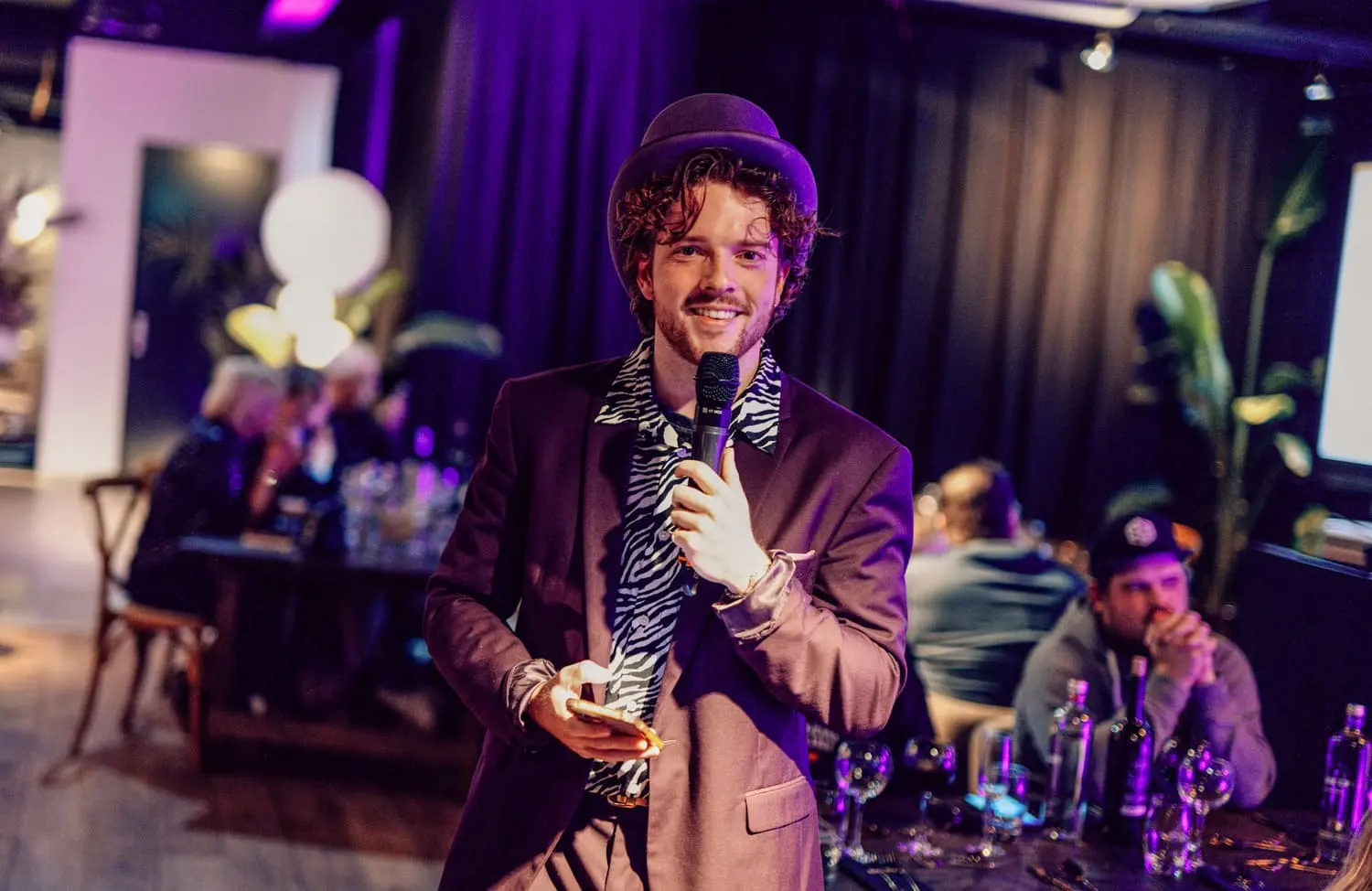 Gastheer in goud jasje houdt microfoon vast tijdens een energieke pubquizshow, omringd door enthousiast publiek. Pubquiz inhuren evenement.