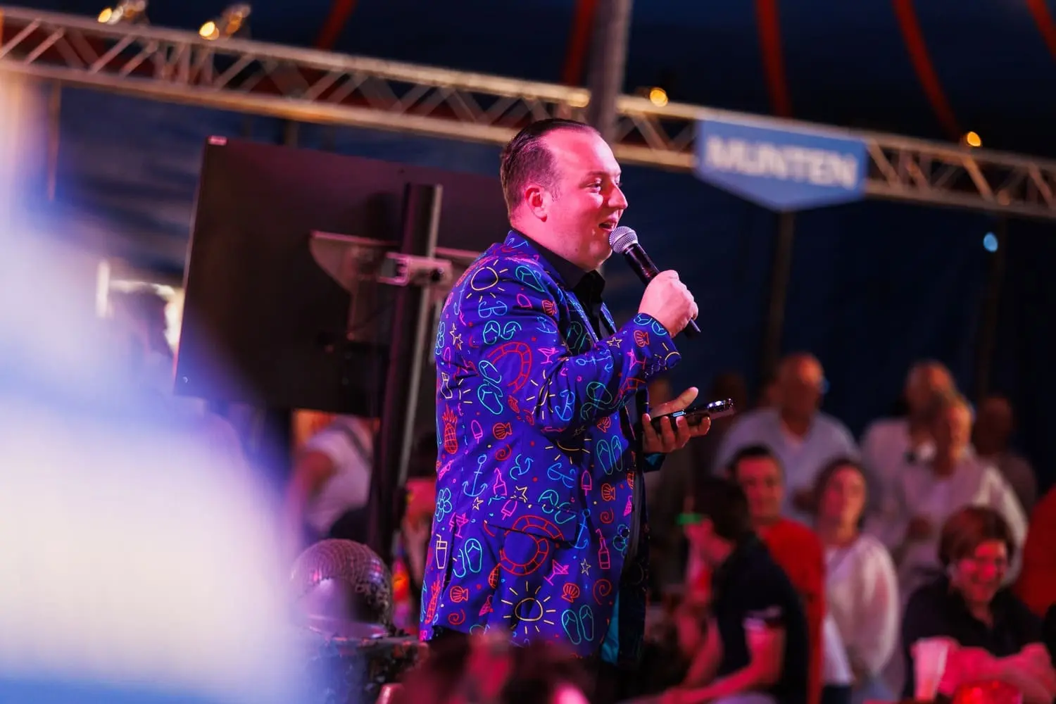 Man met kleurrijk jasje en microfoon presenteert tijdens een Crazy Disco Bingo evenement, met een betrokken publiek en feestelijke sfeer op de achtergrond.