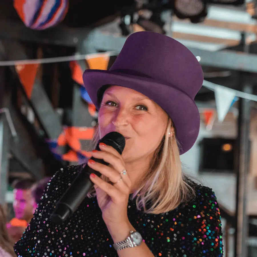 Evenementmanager met paarse hoed en microfoon op een kleurrijk feest - Linda Spaans van QuizX.