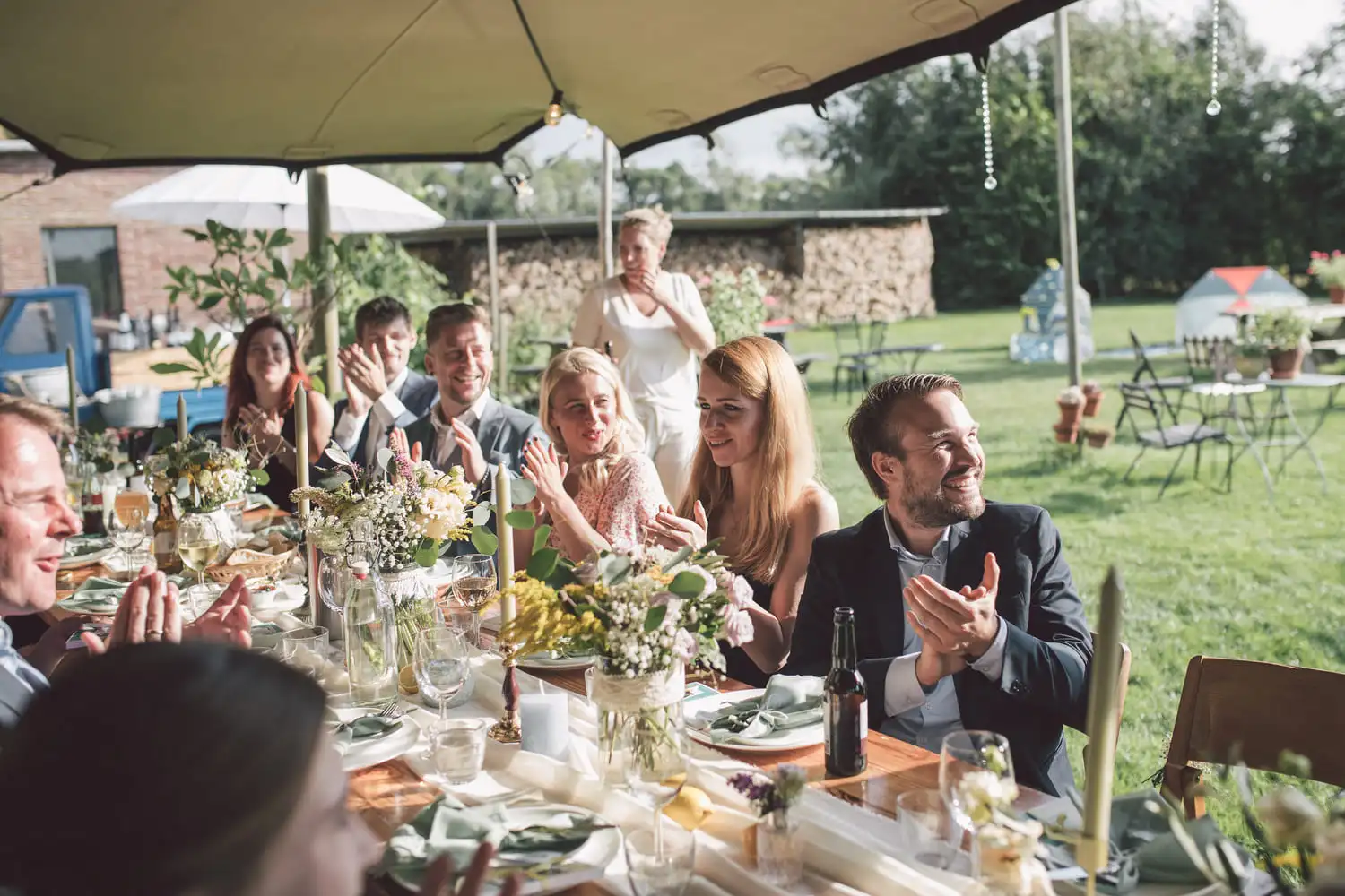 Groep mensen geniet van een feestelijke maaltijd buiten met bloemen en kaarsen op tafel, passend bij een gepersonaliseerde pubquiz sfeer.