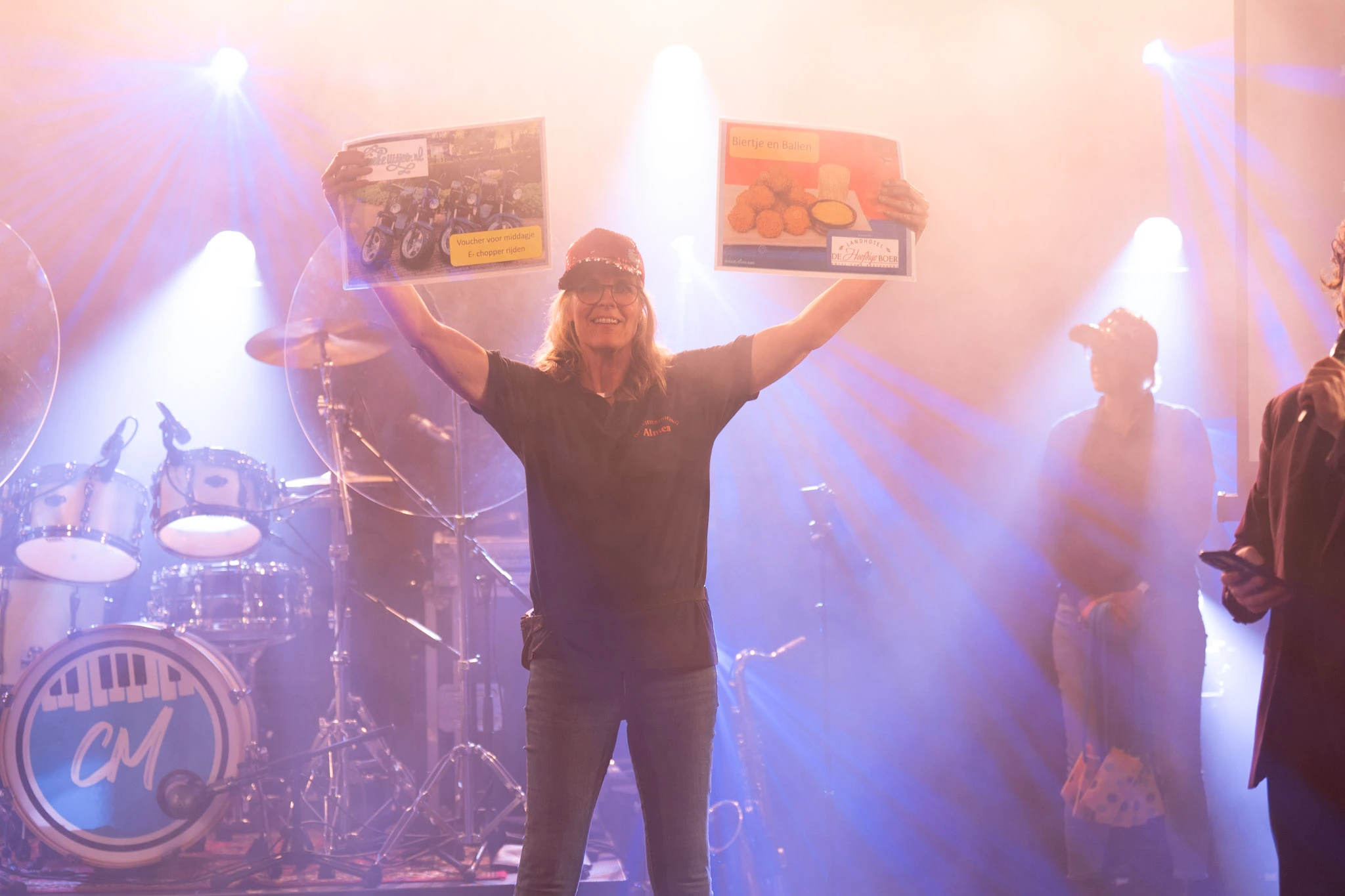 Persoon op podium houdt certificaten omhoog tijdens feestelijk evenement, met kleurrijke lichten en drumstel op de achtergrond. Context: Pubquiz Den Haag.