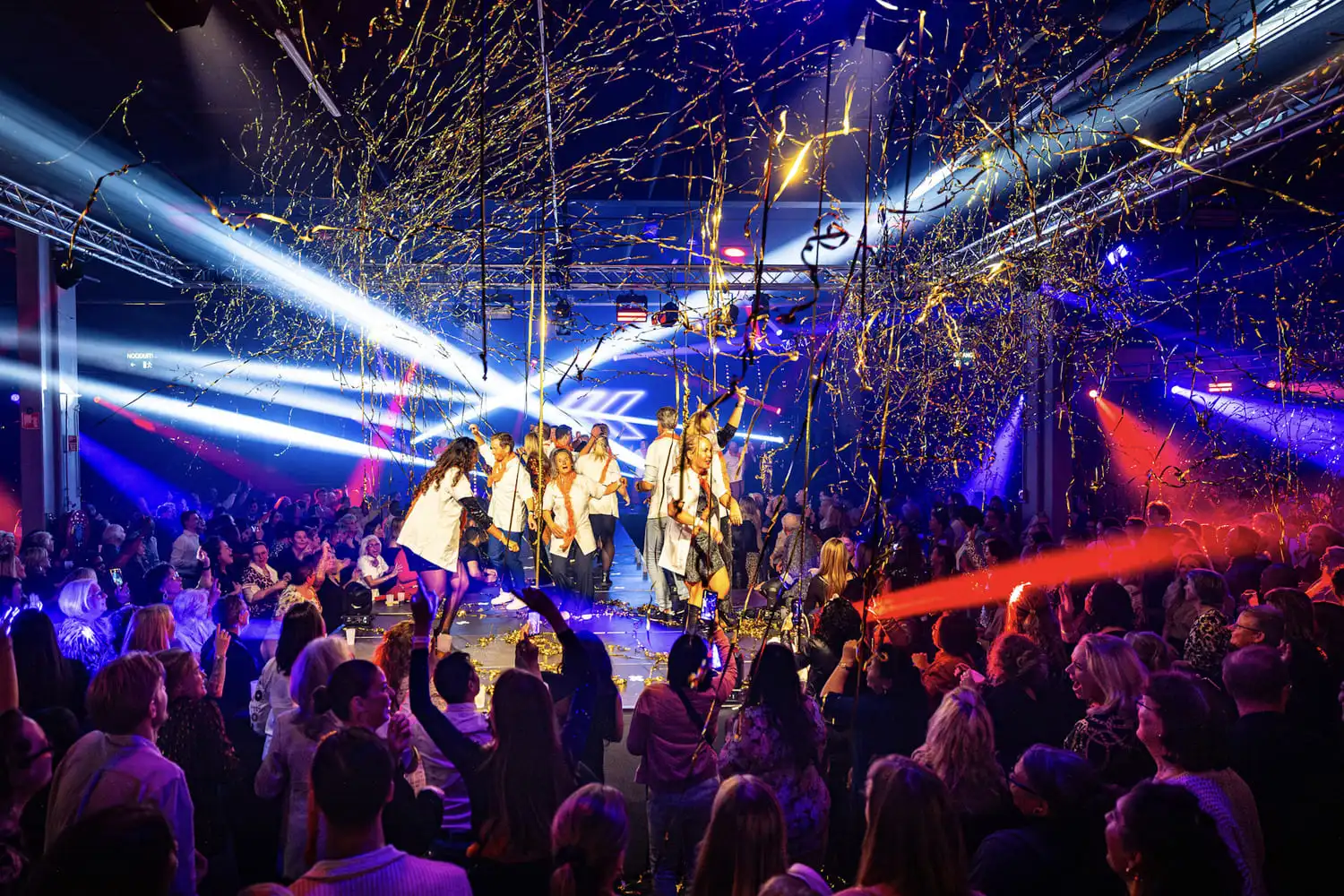 Een levendige en feestelijke scène tijdens een pubquiz op locatie, met mensen die dansen op een podium te midden van kleurrijke lichteffecten en confetti in de lucht.