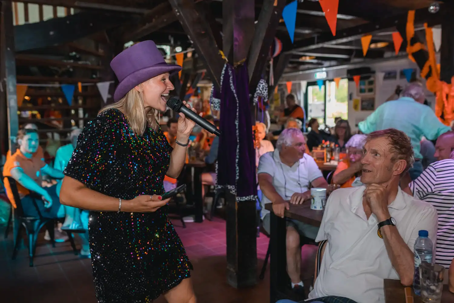 Presentatrice in glinsterende paillettenjurk en paarse hoed tijdens een quizavond bij Ik hou van Holland.