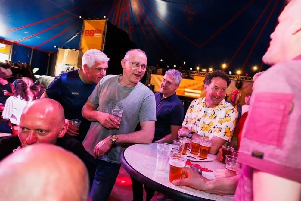 Mensen genieten samen van een gezellige avond in een tent tijdens een gepersonaliseerde pubquiz, omringd door kleurrijke verlichting en bierglazen.