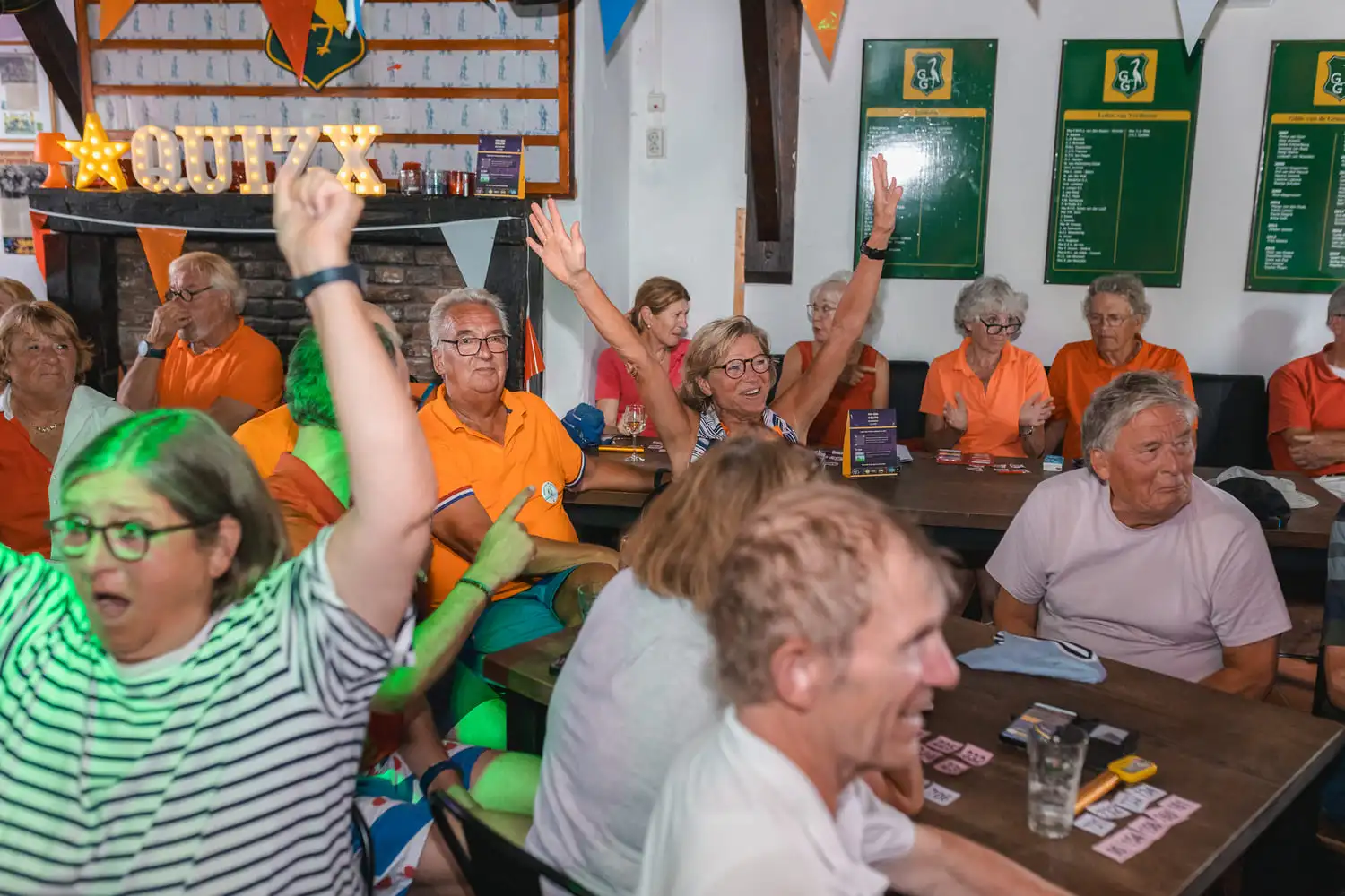 Groep enthousiaste oudere deelnemers aan een 'Ik hou van Holland quiz' in oranje kleding, lachend en juichend in een café met verlichte 'QUIZ' letters.
