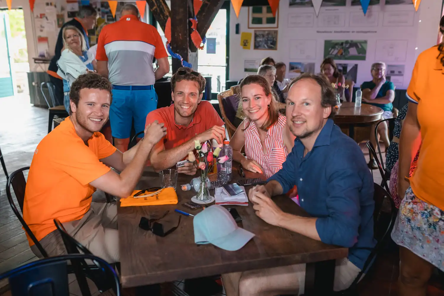 Groep vrienden lachend tijdens een 'Ik hou van Holland quiz', met oranje en blauwe kleding in een feestelijk versierde ruimte.