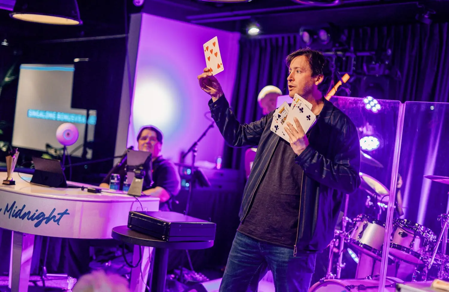 Persoon voert een kaarttruc uit op een feestelijke pubquiz-avond met live muziek en interactieve entertainment in een paarse verlichte omgeving.