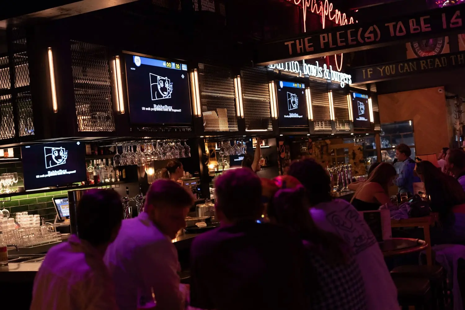 Een levendige bar in Breda met gasten die deelnemen aan een pubquiz terwijl televisieschermen quizvragen tonen.