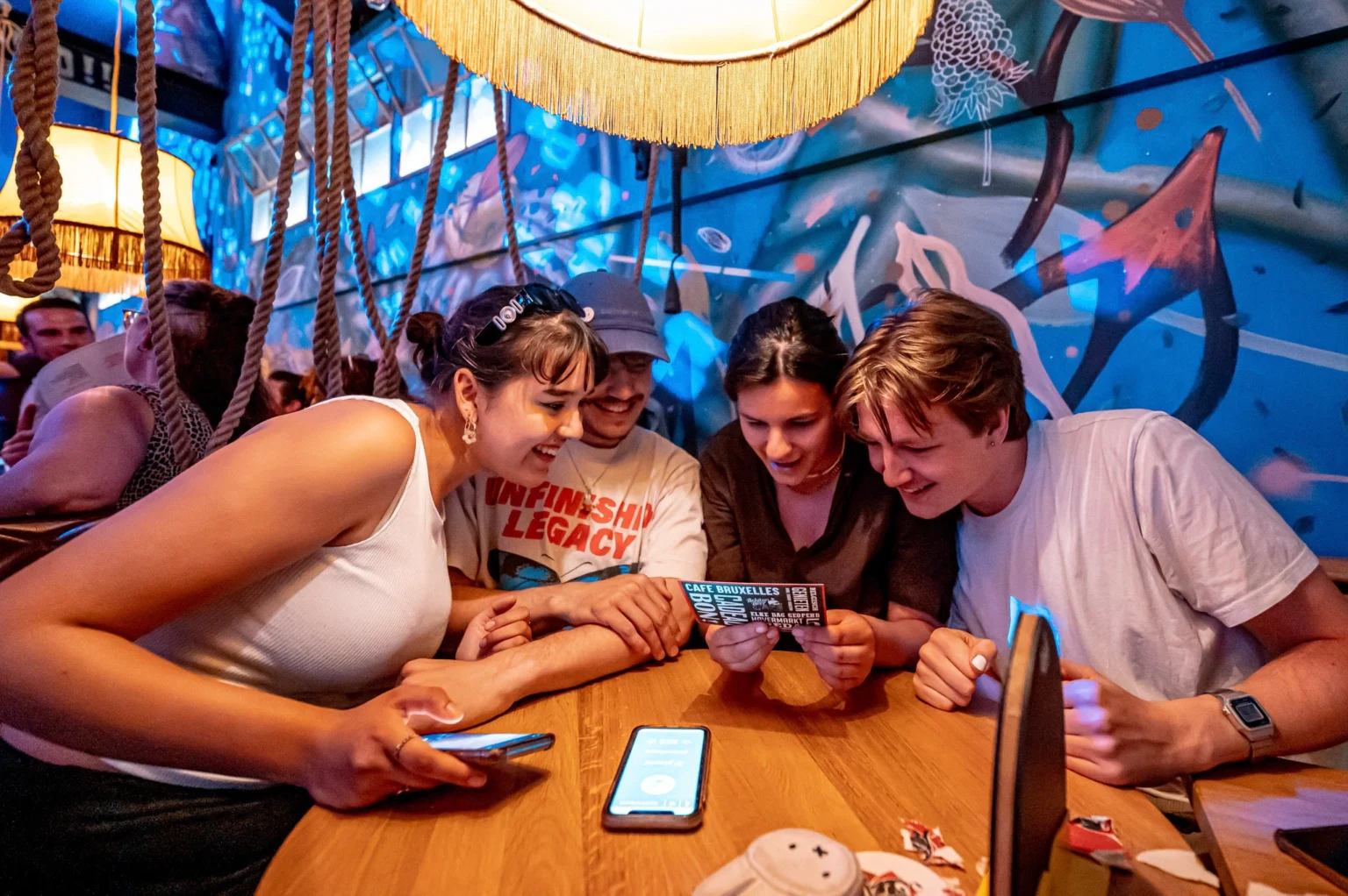 Groep vrienden geniet van een pubquiz in een café in Den Haag, kijkend naar een flyer en een telefoon op een houten tafel in een sfeervolle, artistiek ingerichte ruimte.