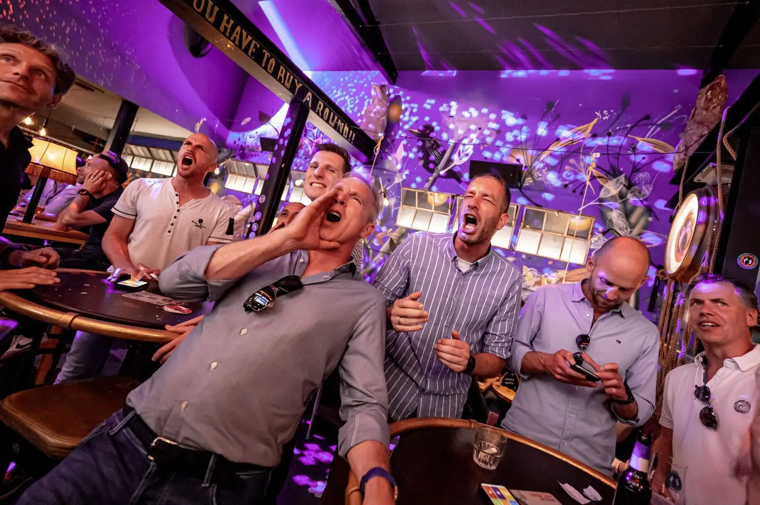 Groep mannen neemt enthousiast deel aan een pubquiz in een bar met feestelijke paarse verlichting.