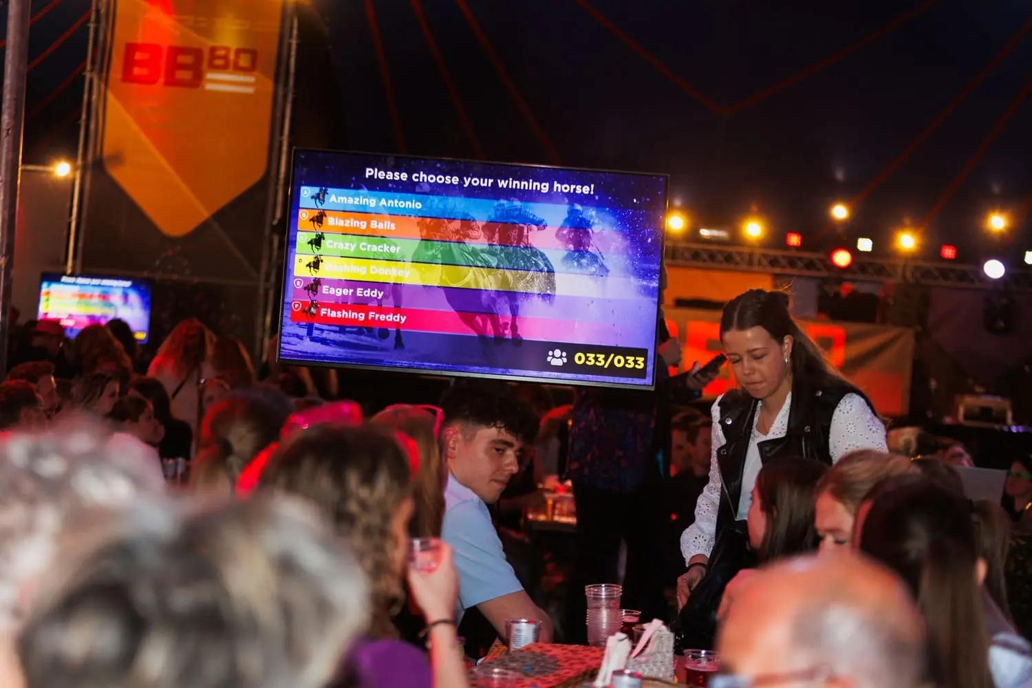 Een drukbezocht pubquiz evenement met een groot scherm dat een interactieve paardenrace toont. Mensen zitten aan tafels met drankjes, genietend van de gepersonaliseerde pubquiz ervaring.