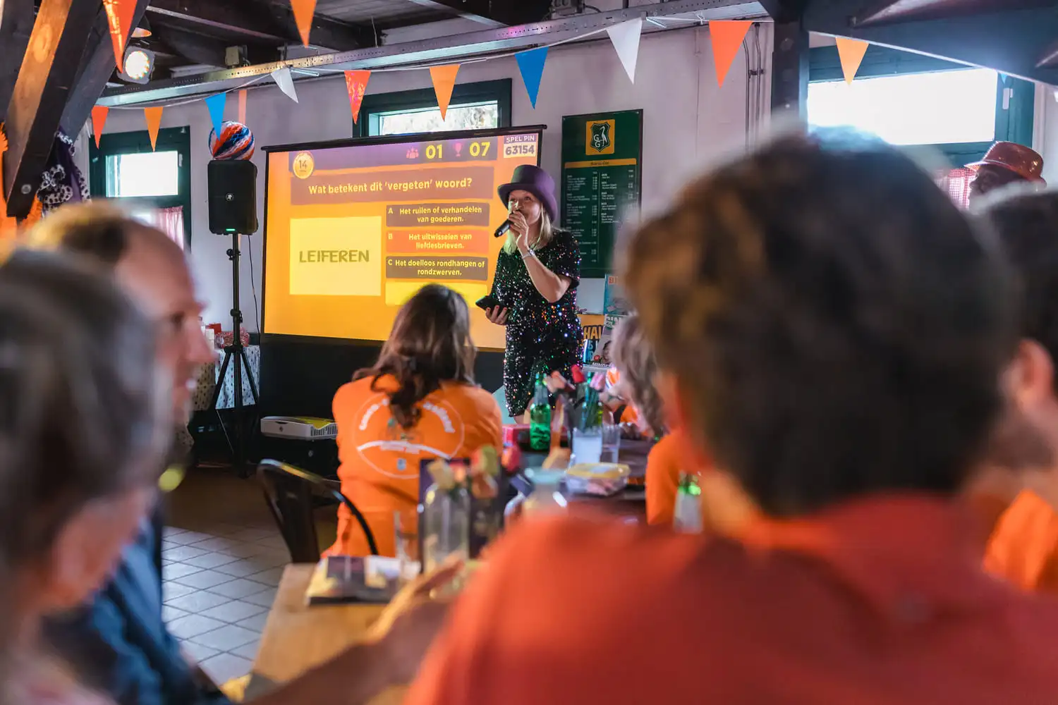 Interactieve pubquiz waar een vrouw met microfoon de vraag 'Wat betekent het vergeten woord leiferen?' stelt in een feestelijke setting.