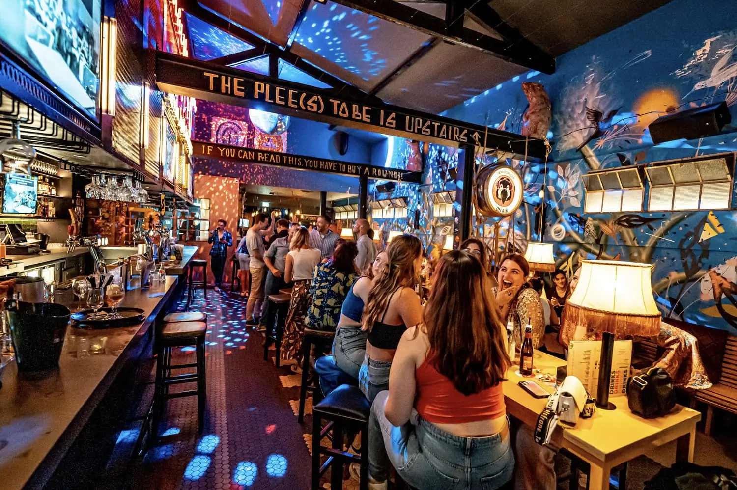 Een levendige baromgeving met mensen die genieten van een pubquiz op locatie, omgeven door kleurrijke lichteffecten en muurschilderingen.