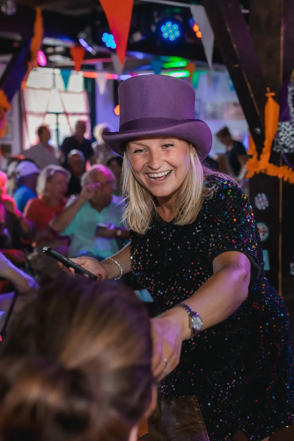 Vrouw in paarse hoed lacht tijdens een pubquiz op locatie in een feestelijk gedecoreerde ruimte.